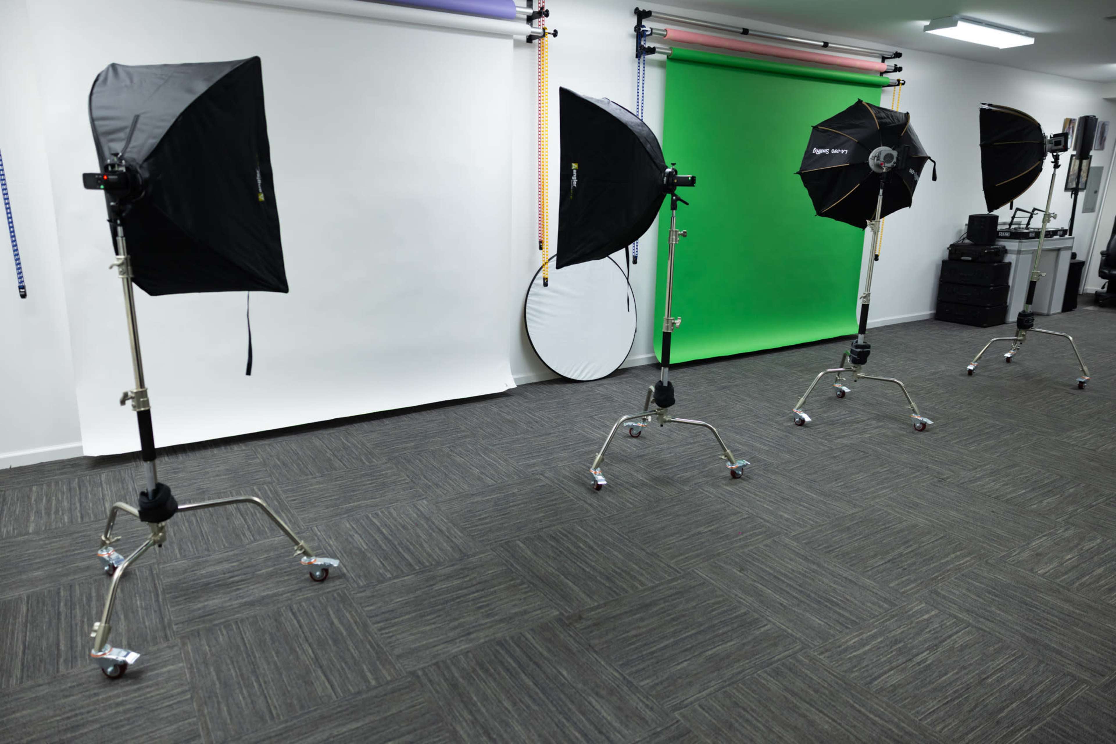 The image shows a photography studio with four light stands equipped with softboxes, positioned on a flat, dark floor, and a green screen backdrop on one side.
