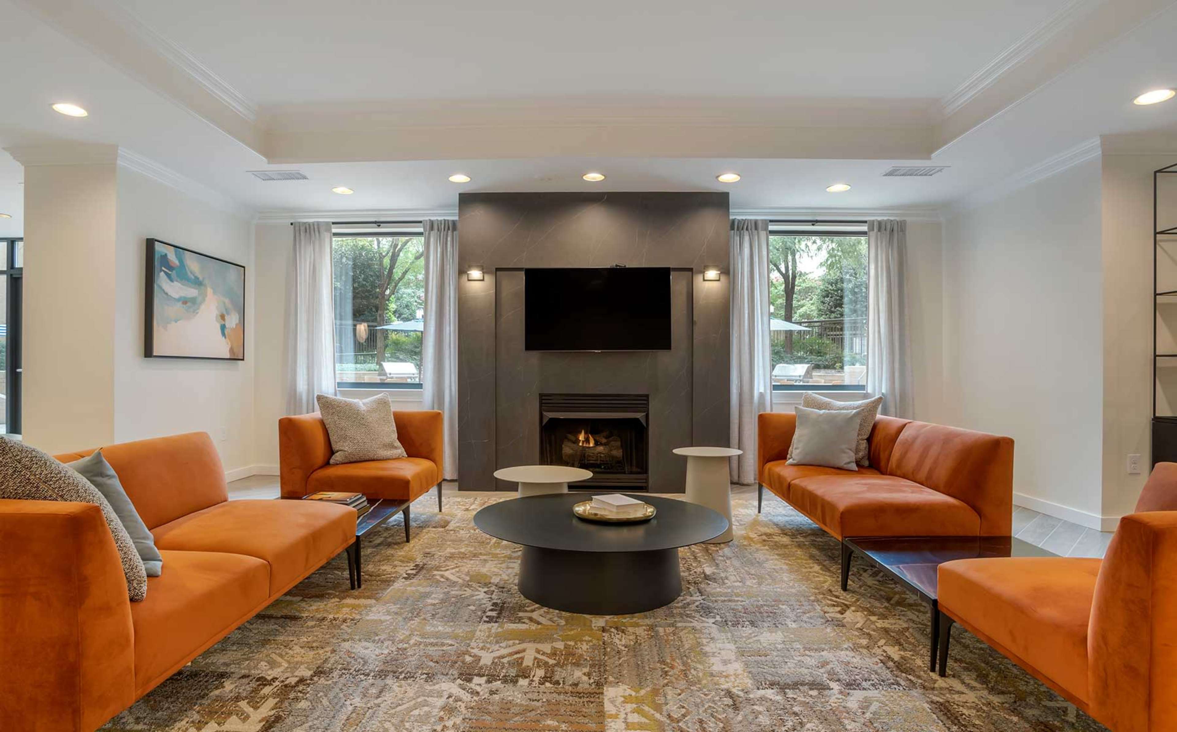 The image shows a modern living room with orange sofas arranged around a central table, a fireplace, and large windows allowing light to enter.