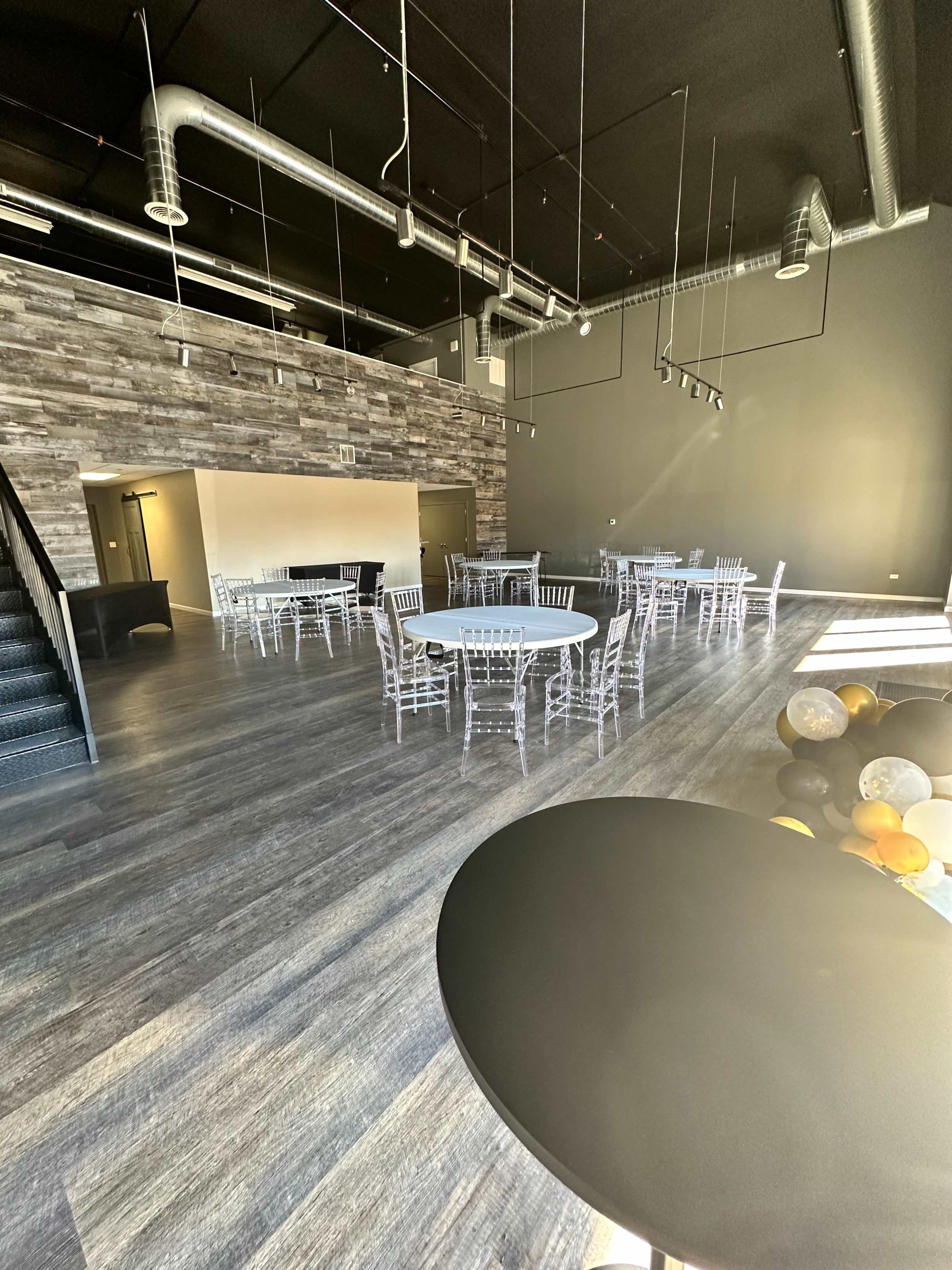 The image shows a spacious event hall with several round tables surrounded by transparent chairs, and a staircase leading to an upper level.