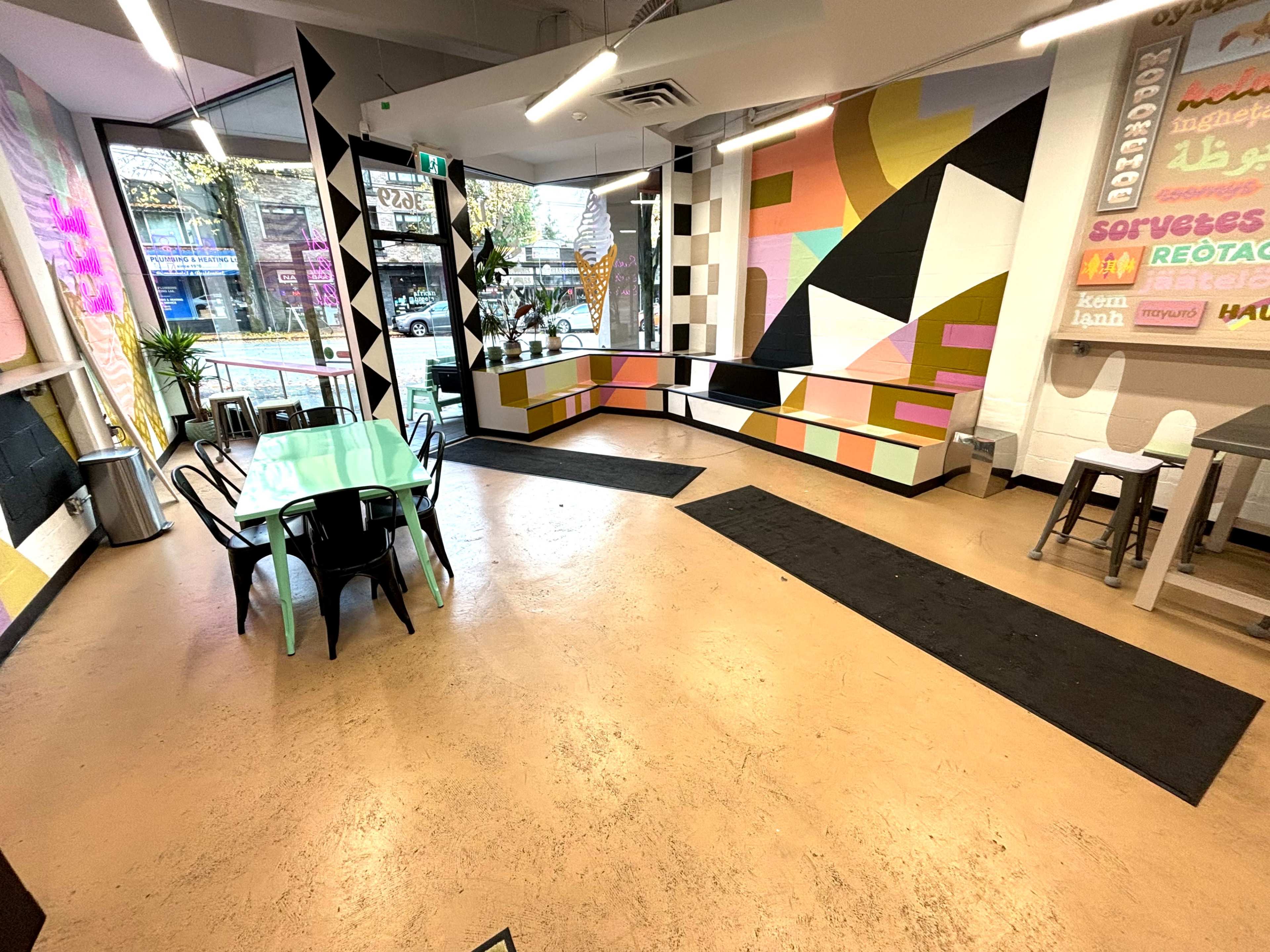 The image shows a modern café interior featuring colorful geometric wall art, a green table with black chairs, and a large window allowing natural light to enter.