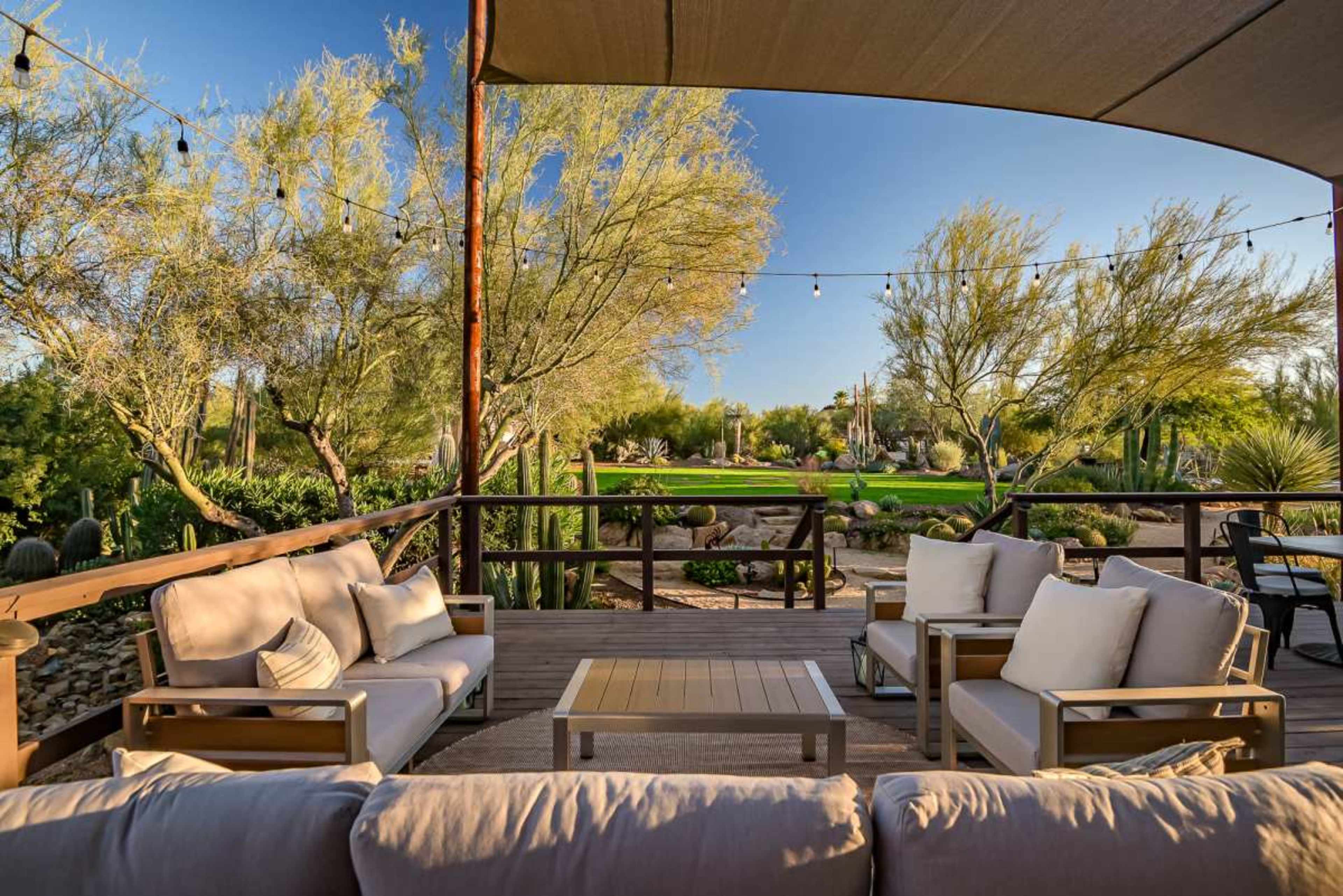 The image shows a patio area with comfortable seating, surrounded by desert plants and cacti, and features overhead string lights.
