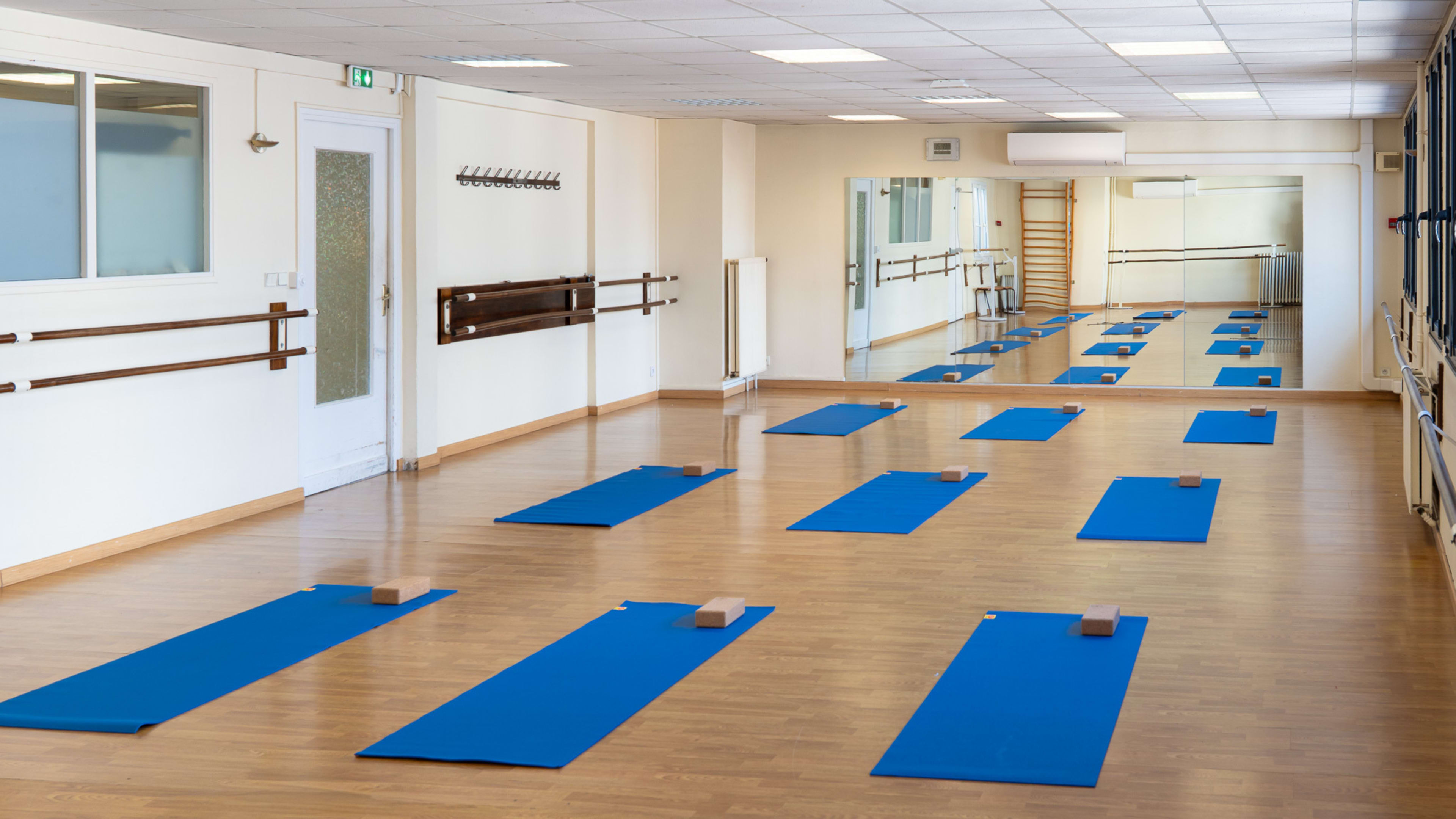 The image shows a yoga studio with several blue mats arranged evenly on a wooden floor, along with a mirrored wall and barre fixtures.