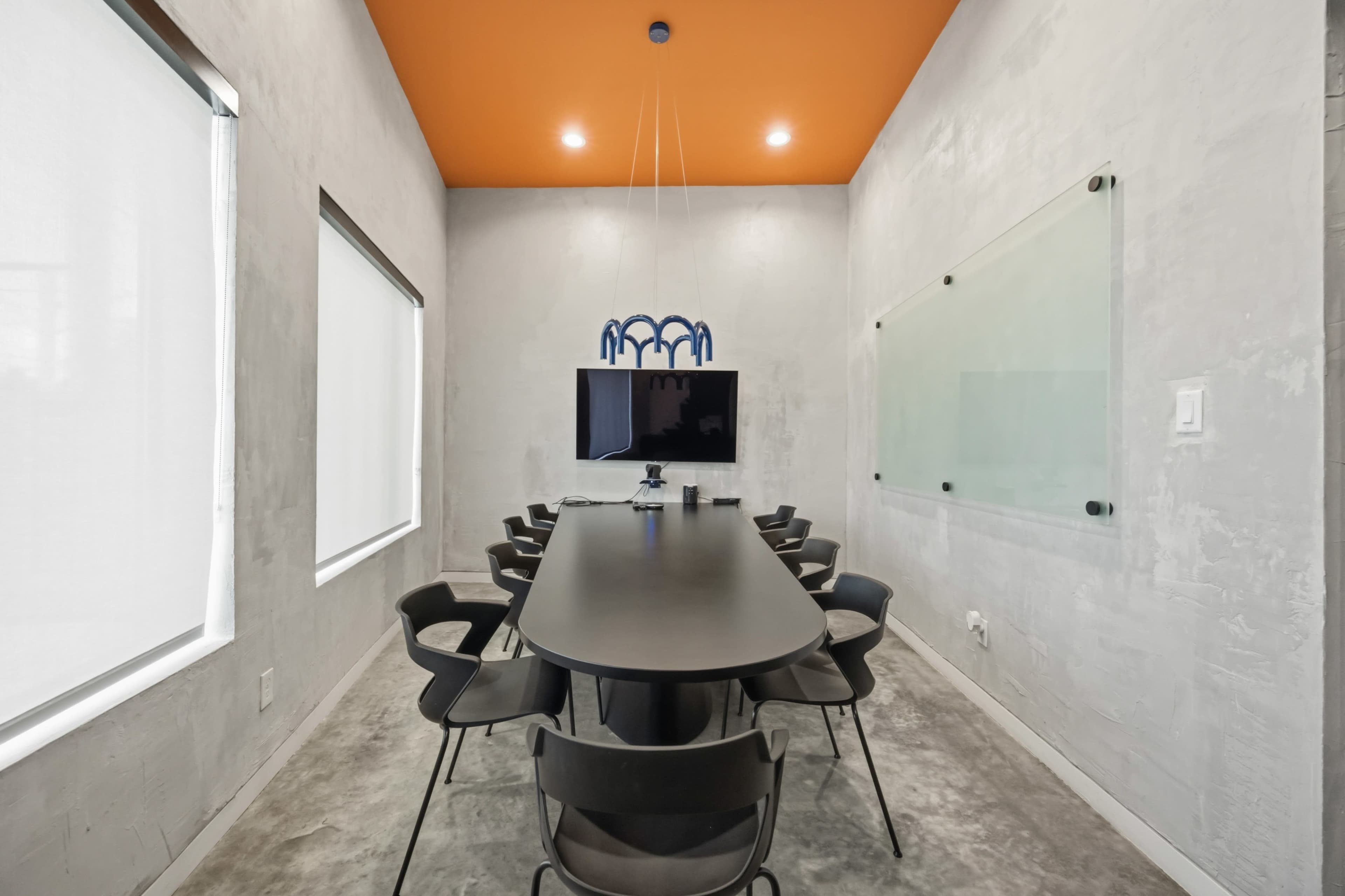 A modern conference room features a long black table, black chairs, a wall-mounted screen, and concrete walls with an orange ceiling.
