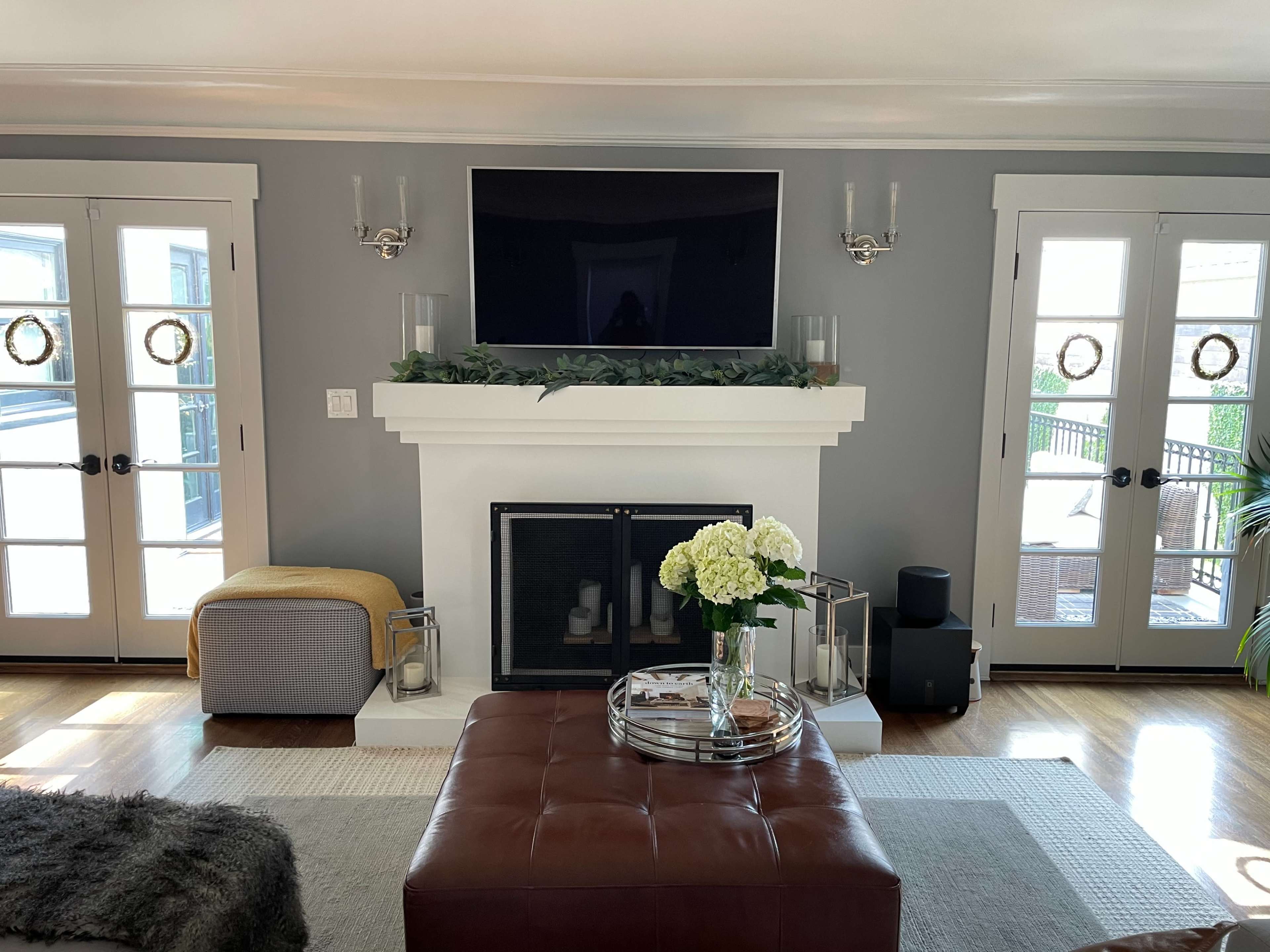 A living room with a fireplace, a mounted television above it, and two sets of double doors leading to an outdoor area, decorated with a floral arrangement on a coffee table.