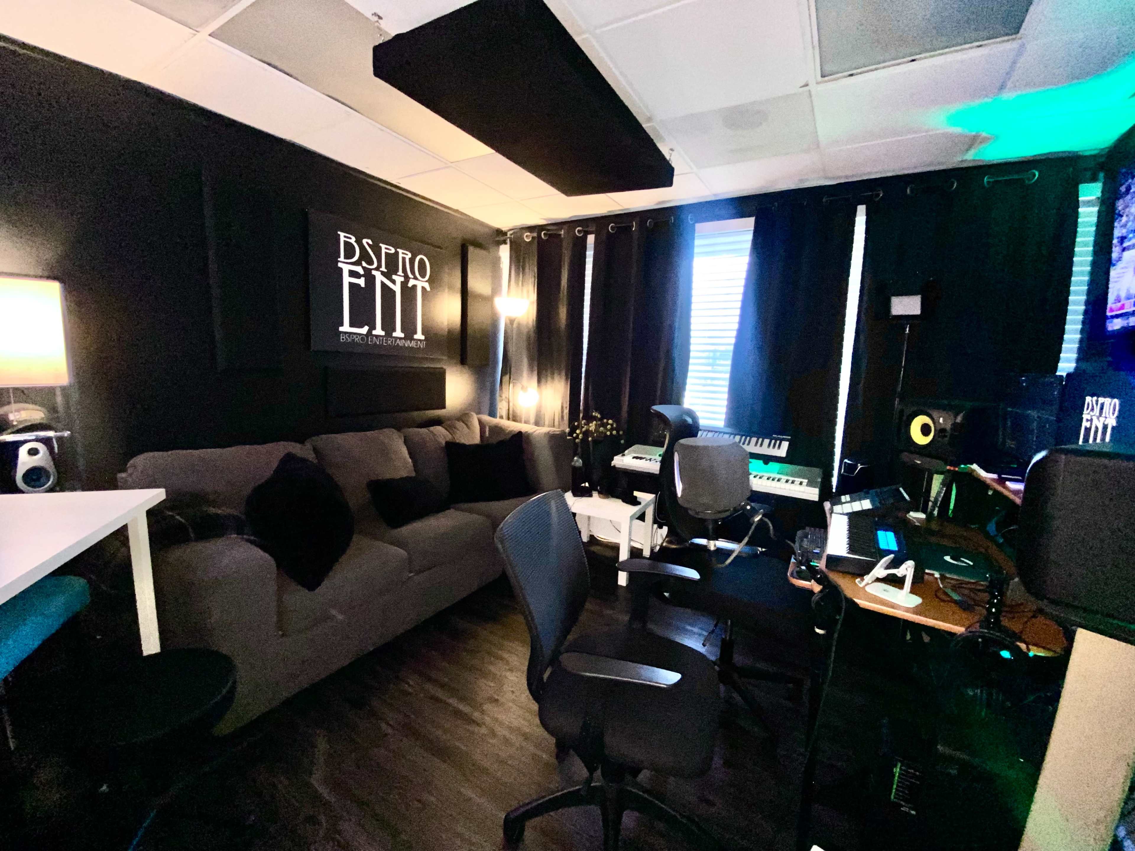 The image shows a modern music studio with a dark color scheme, featuring a couch, multiple chairs, a keyboard, and various audio equipment.