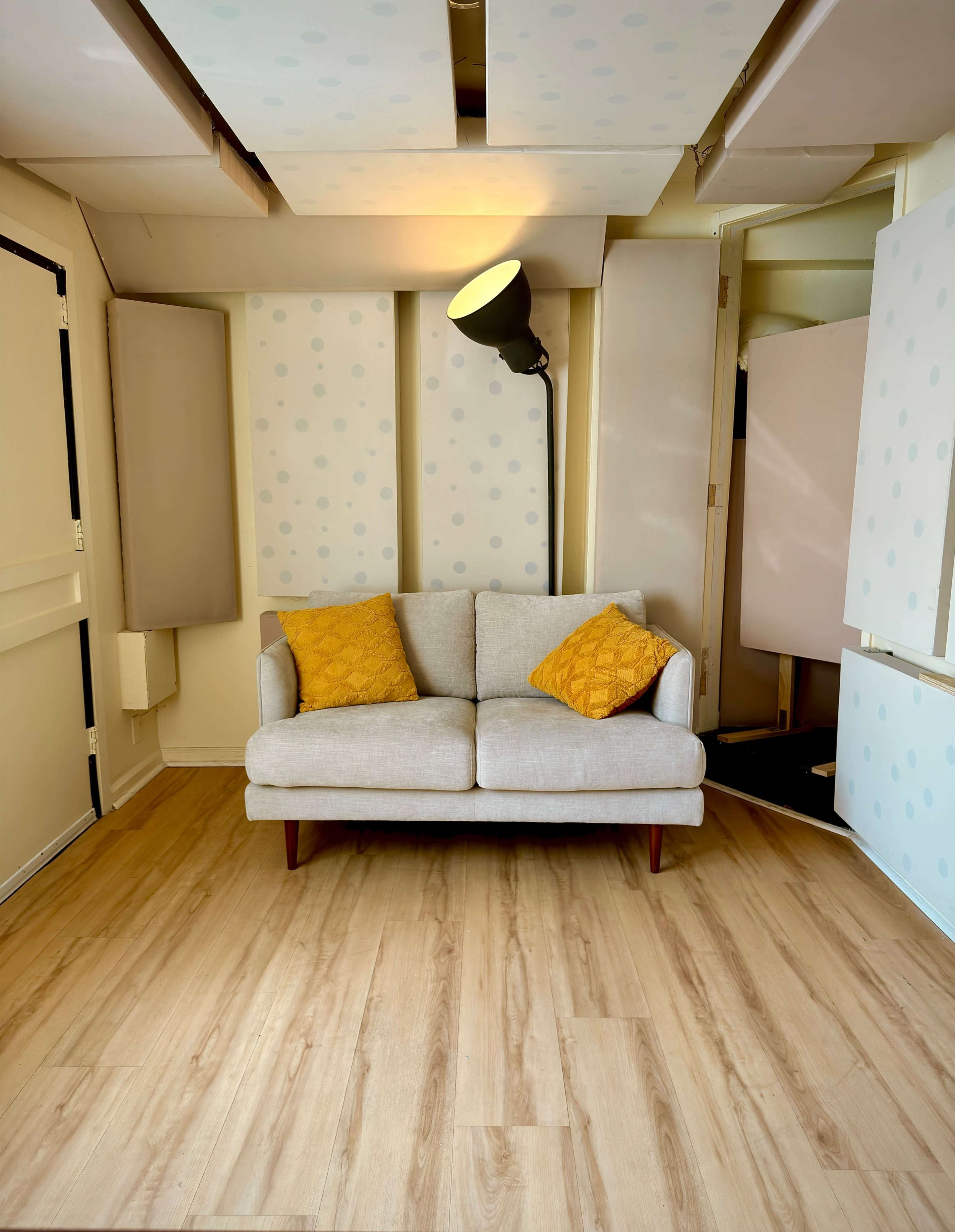 The image shows a simple living space with a light-colored sofa and two yellow throw pillows, positioned against pastel-colored wall panels and illuminated by a floor lamp.