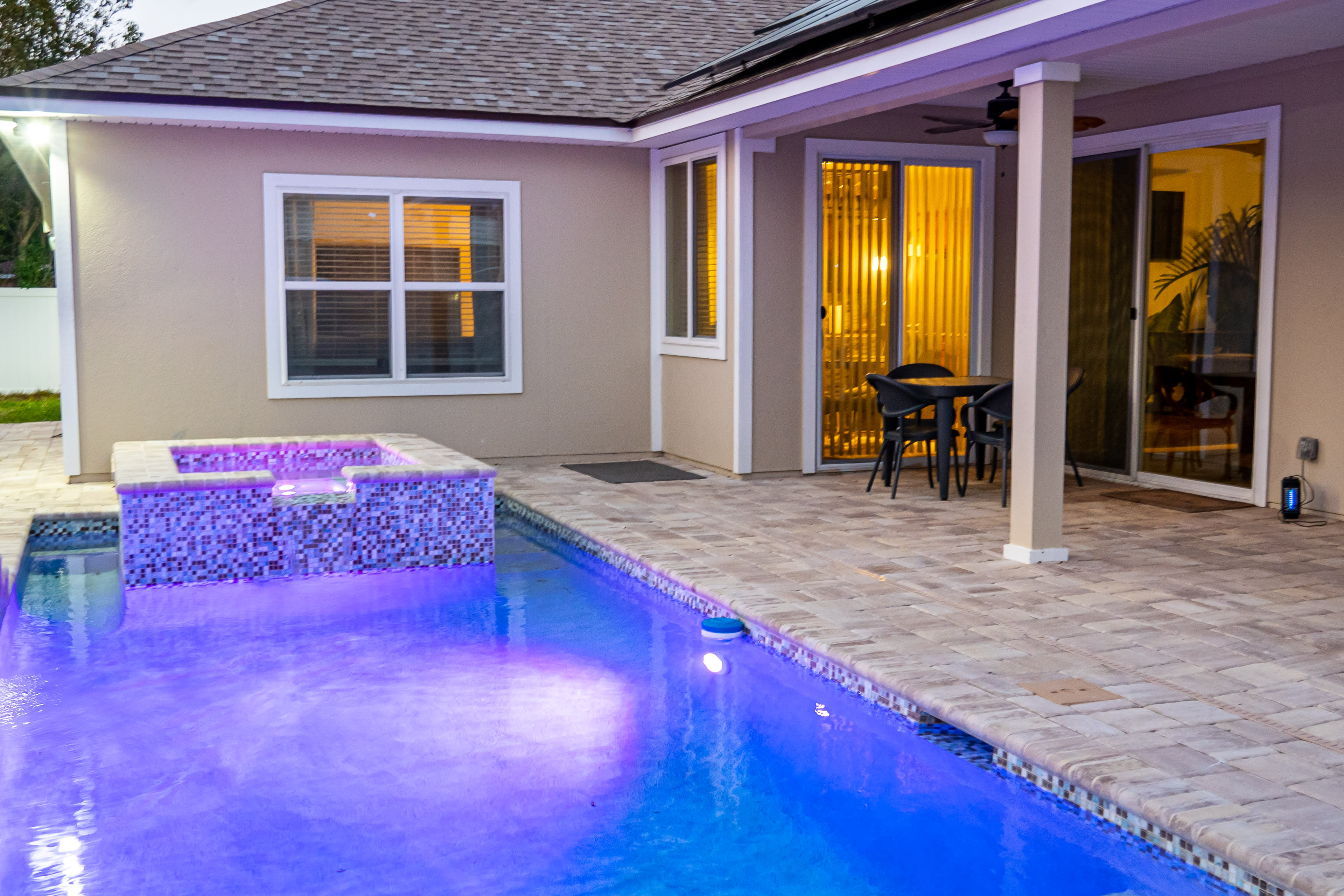 A backyard area with a swimming pool featuring a spa, surrounded by tiled flooring and a set of outdoor furniture under a covered patio.