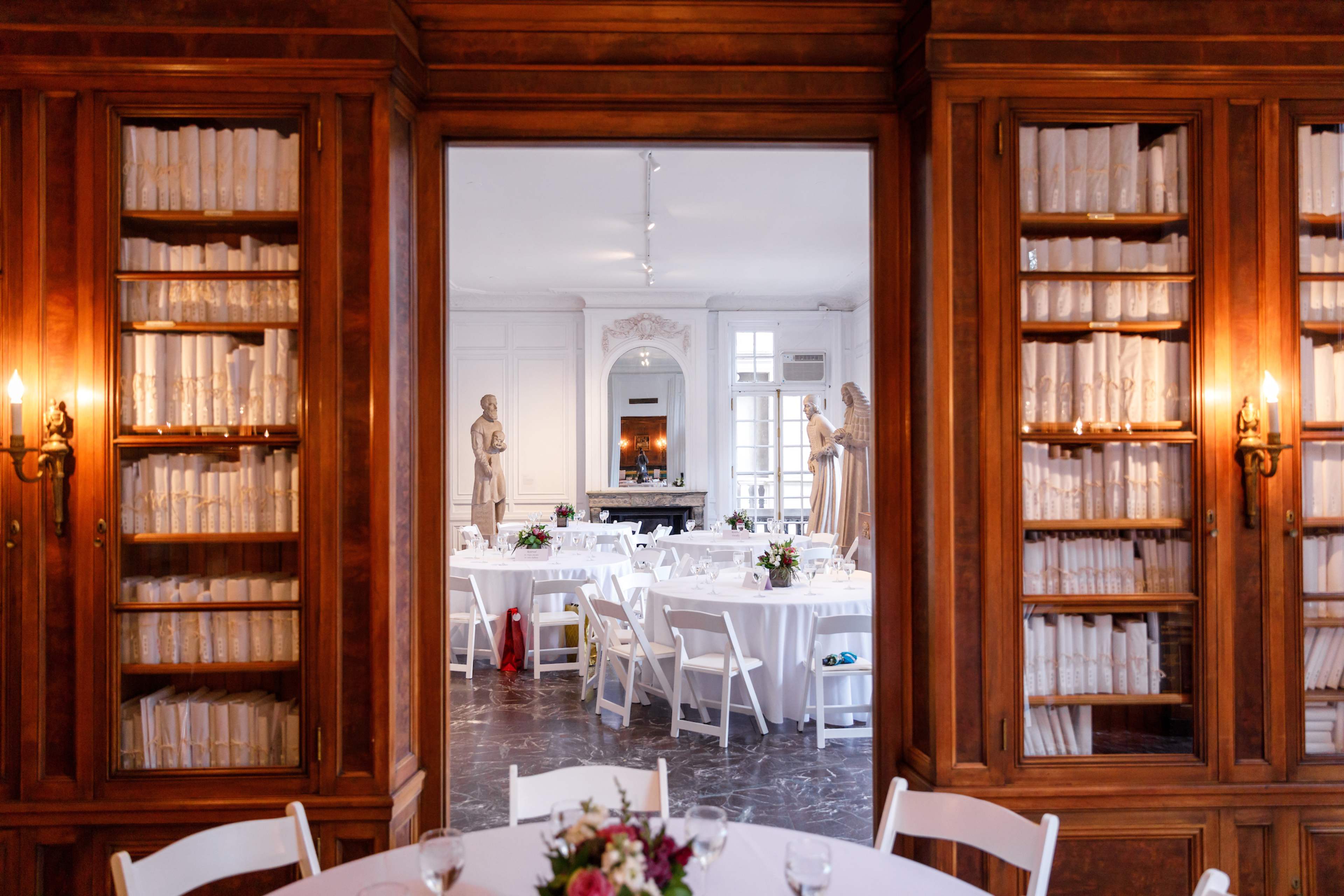 A elegantly set dining room with round tables and white chairs, framed by wooden bookshelves filled with books and flanked by statues.