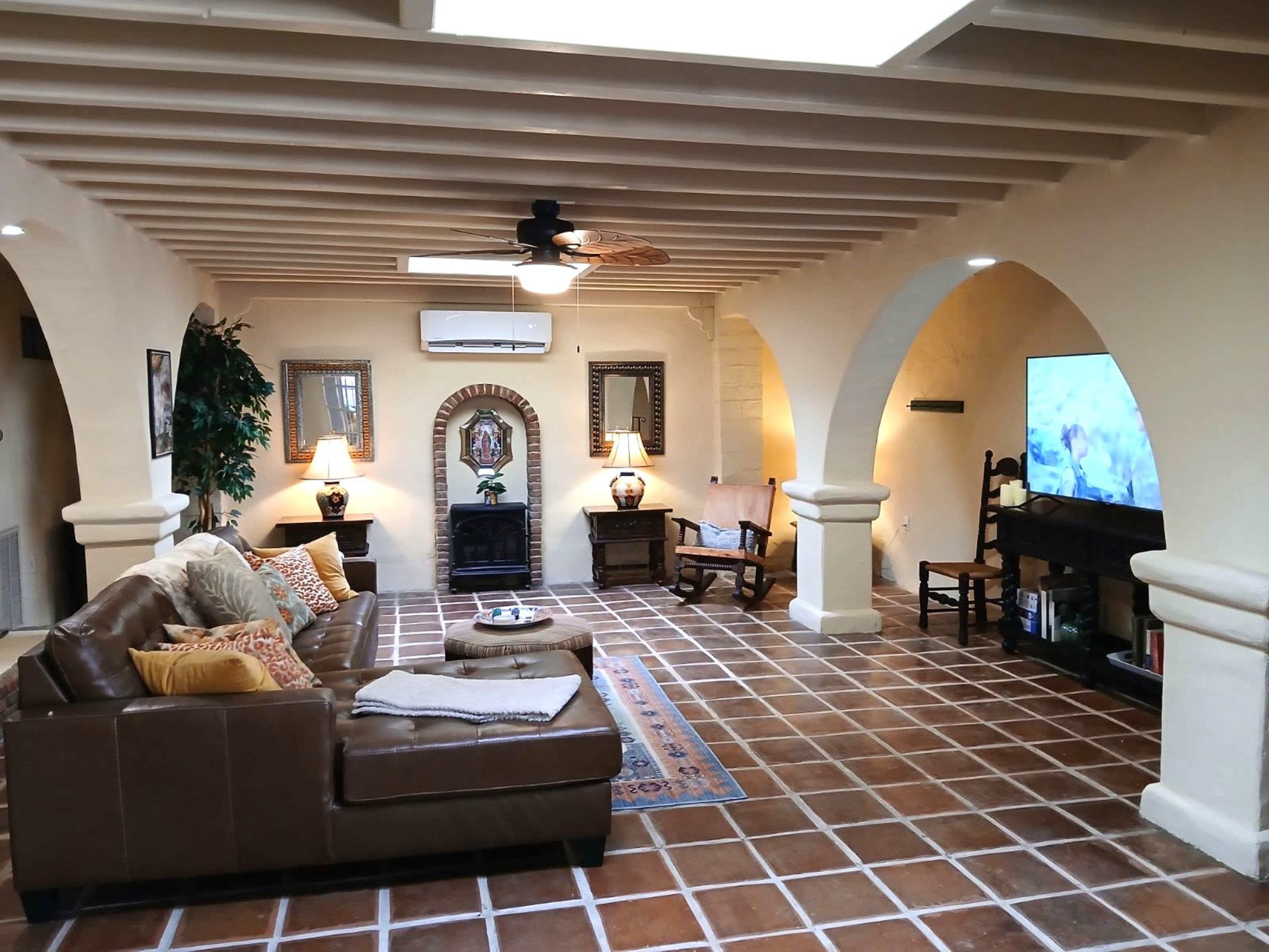 A spacious living room with a brown leather sofa, tile flooring, and archways leading to different areas, featuring a television and a small decorative table.