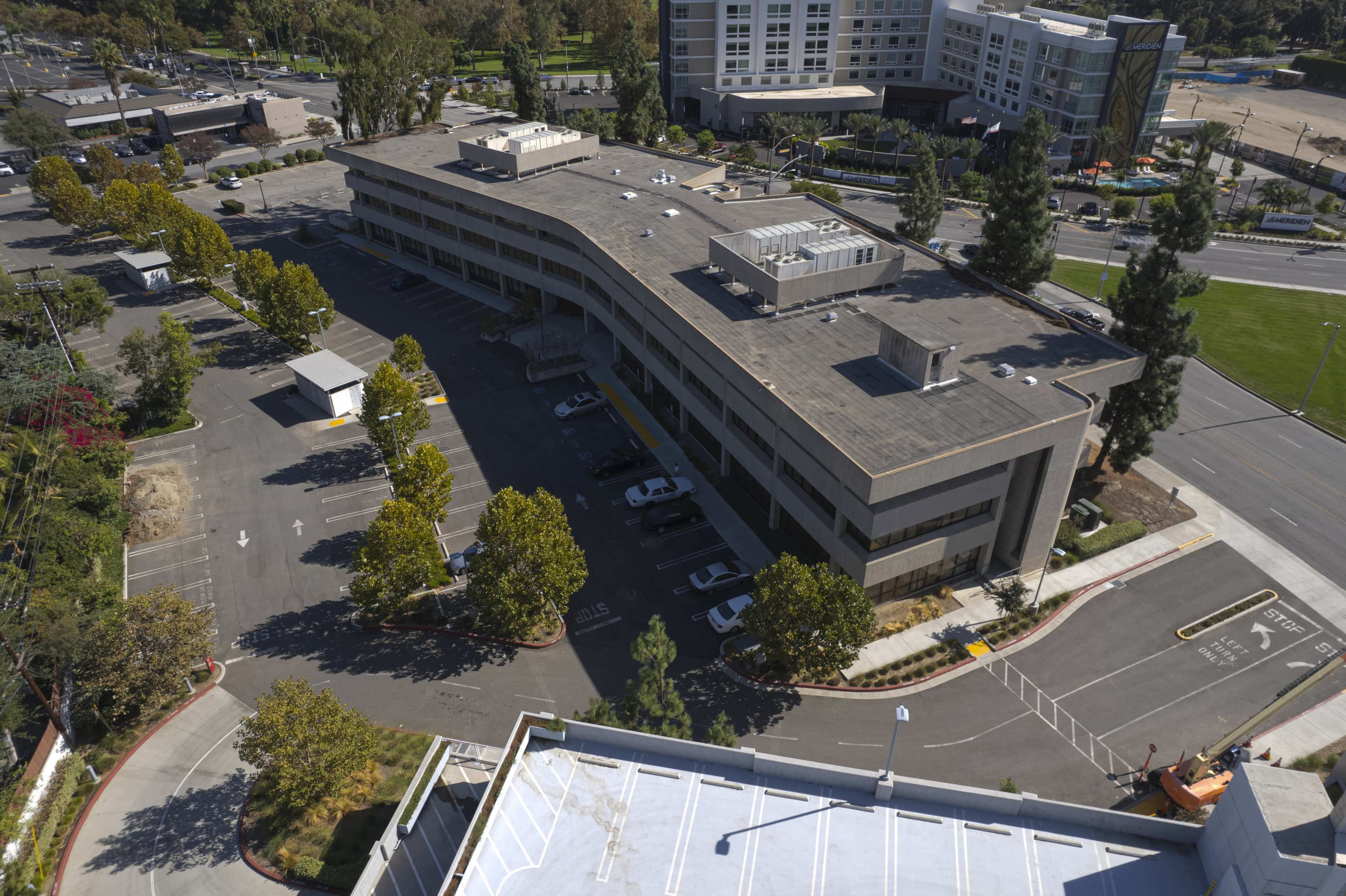 A modern, multi-story building with a parking lot surrounded by trees and adjacent streets in an urban area.