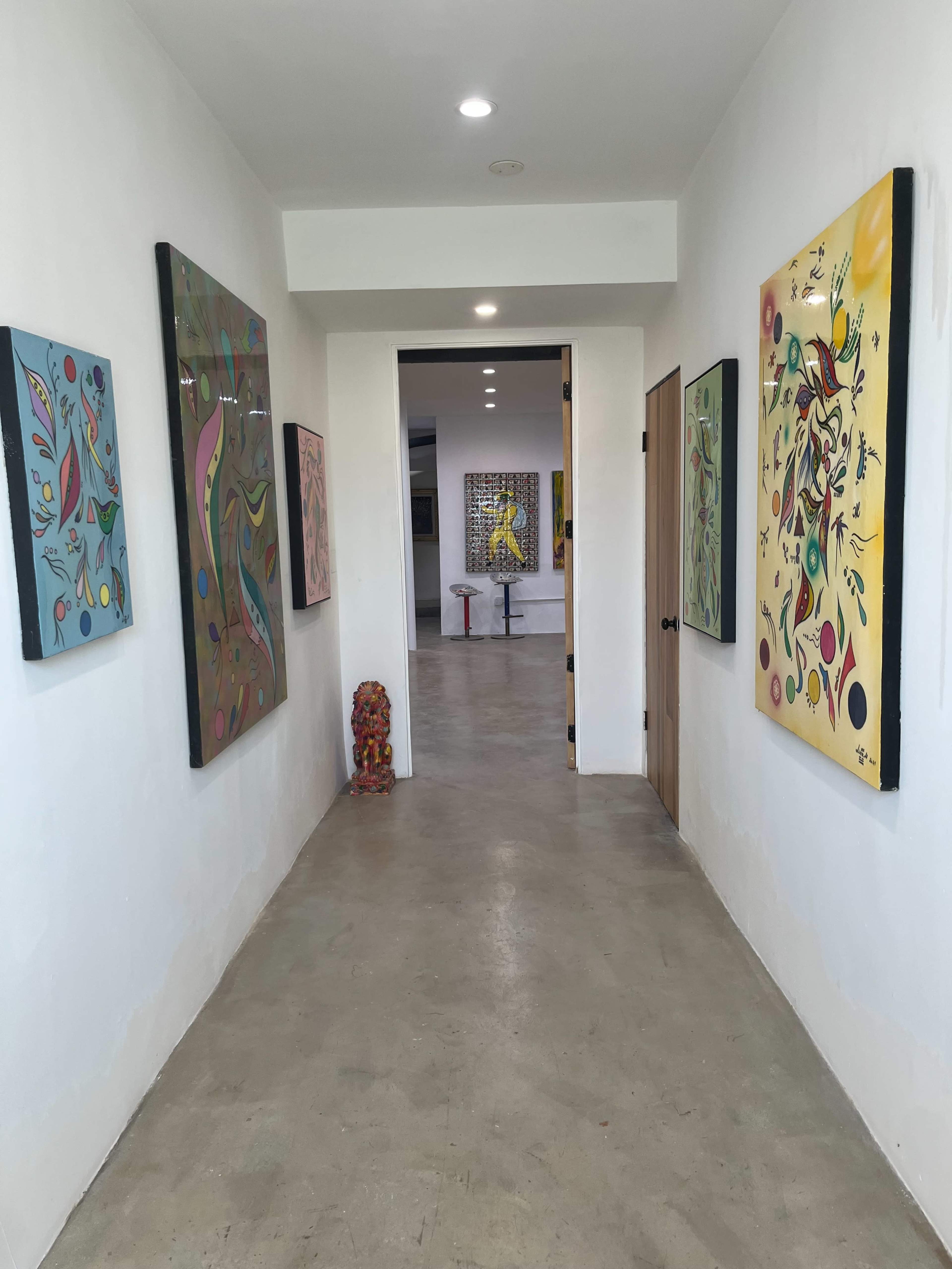 The image shows a hallway lined with colorful artwork on the walls, leading to a brightly lit room with additional art pieces visible at the end.