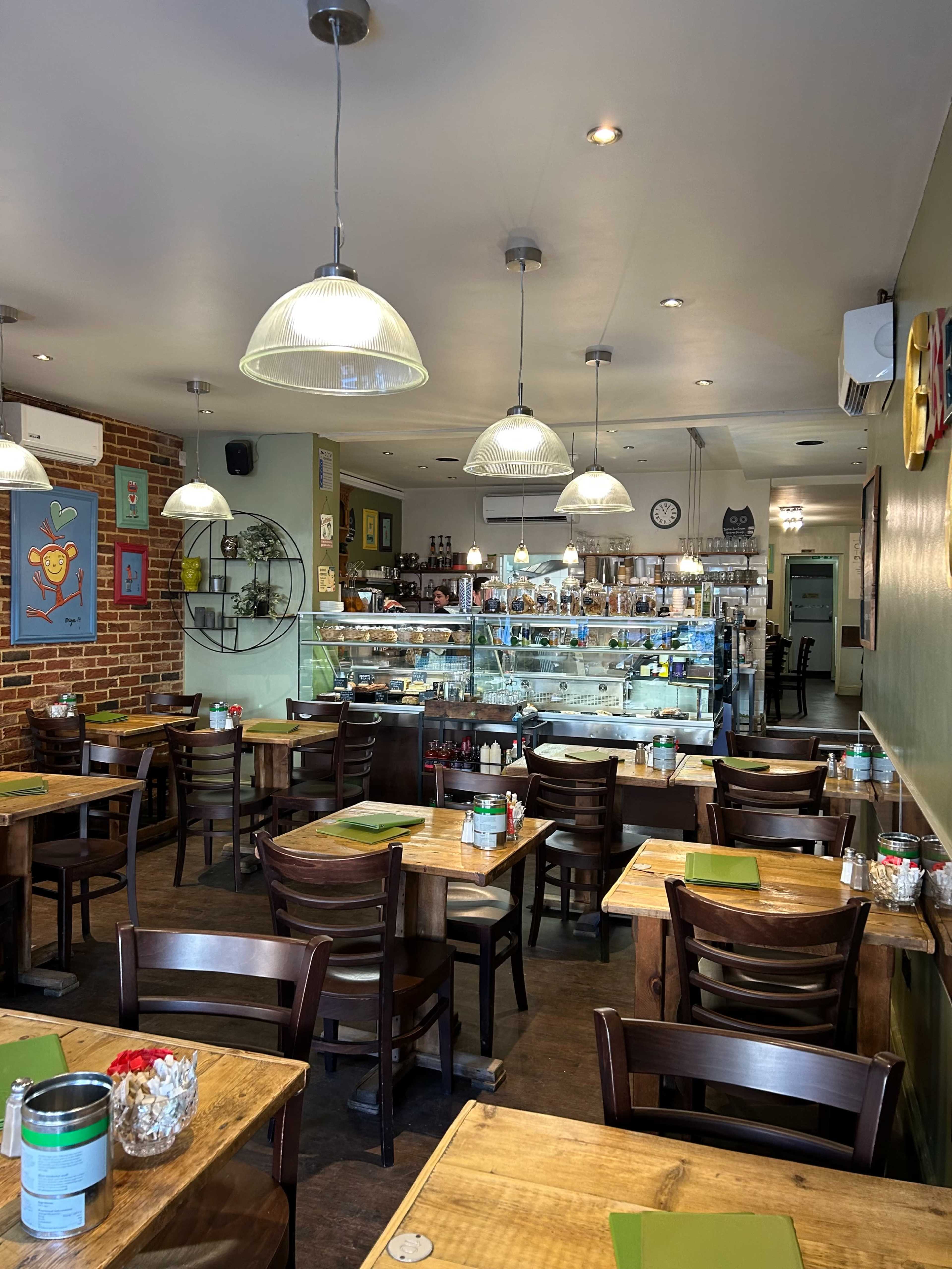 A cozy restaurant interior with wooden tables, green menus, and a display case filled with food, illuminated by pendant lights.