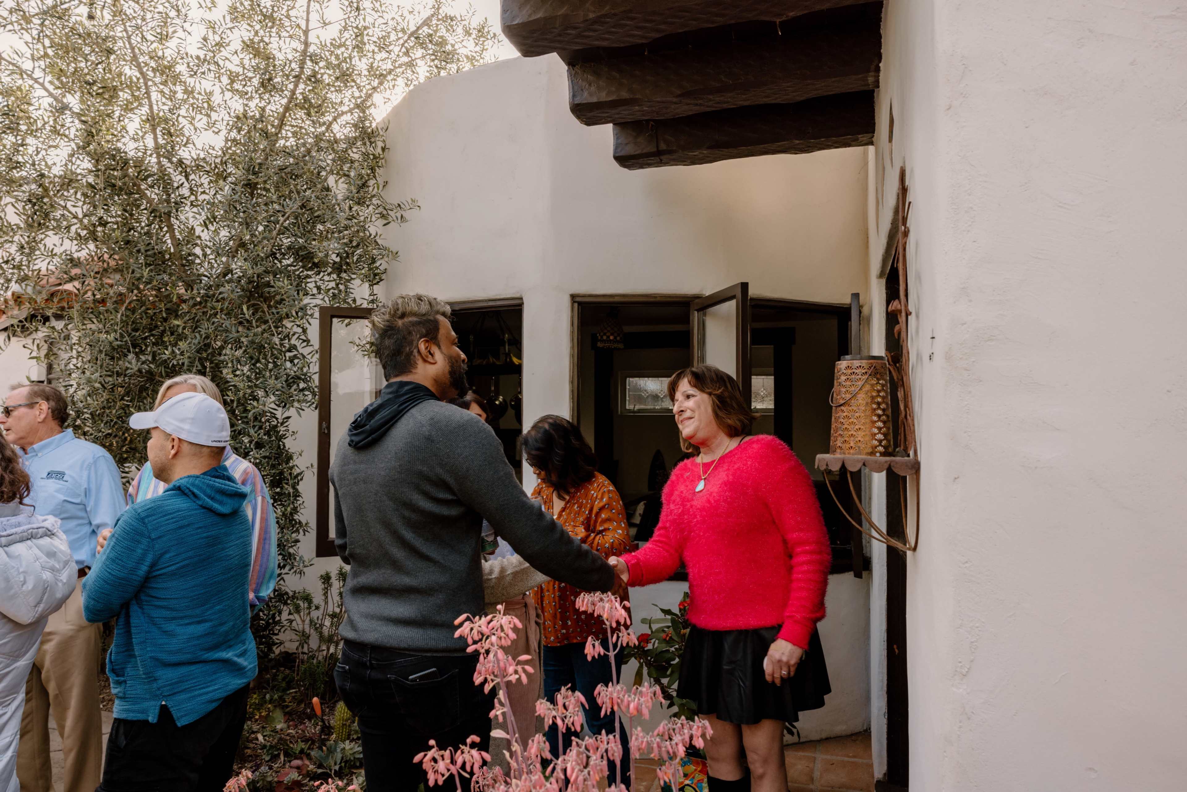 A group of people gathers outside a white building with open doors and a courtyard lined with plants.