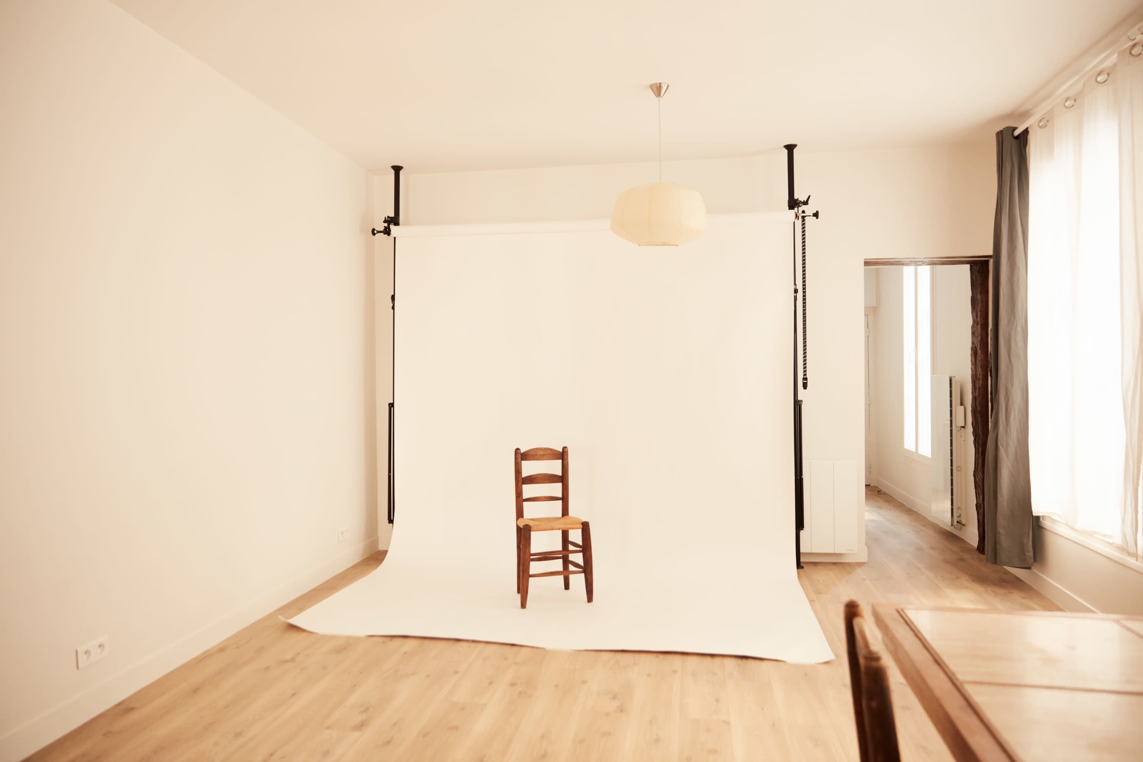 A simple room features a wooden chair placed in front of a white backdrop with soft lighting.