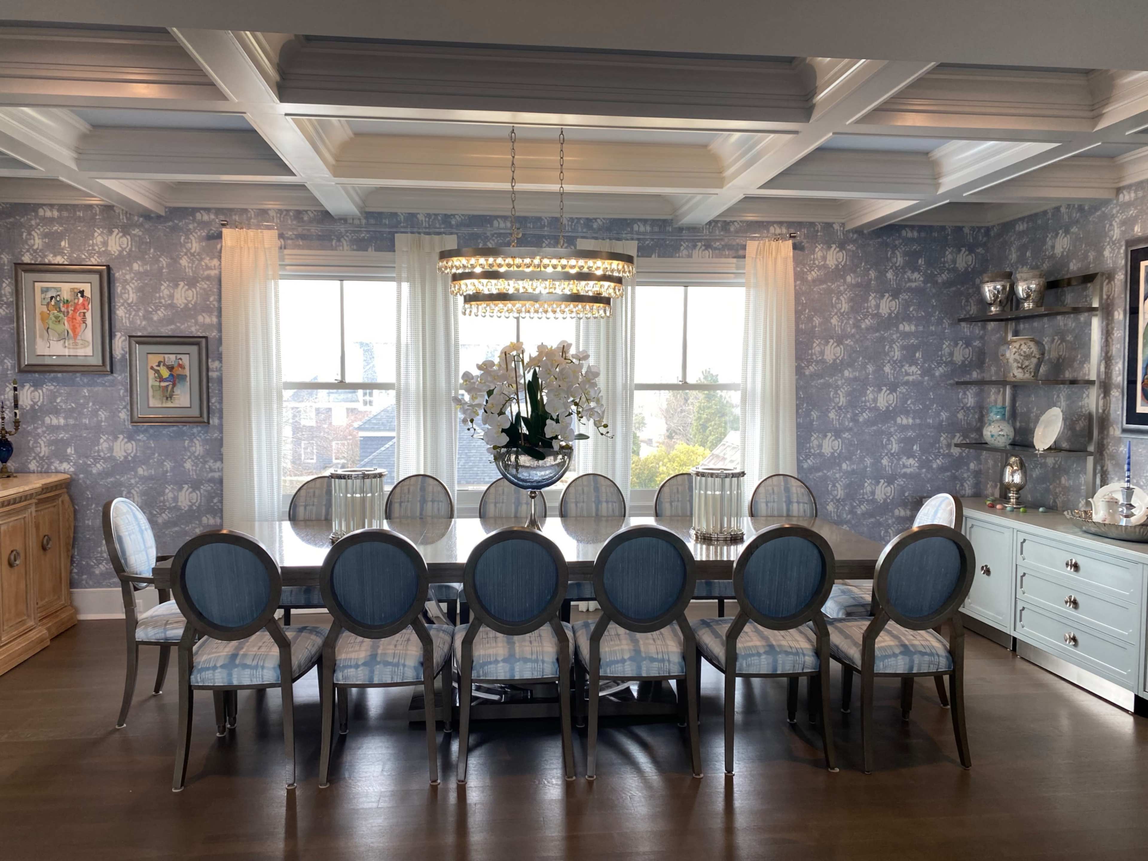 A spacious dining room features a large table surrounded by twelve upholstered chairs, with floral wallpaper and a chandelier overhead.