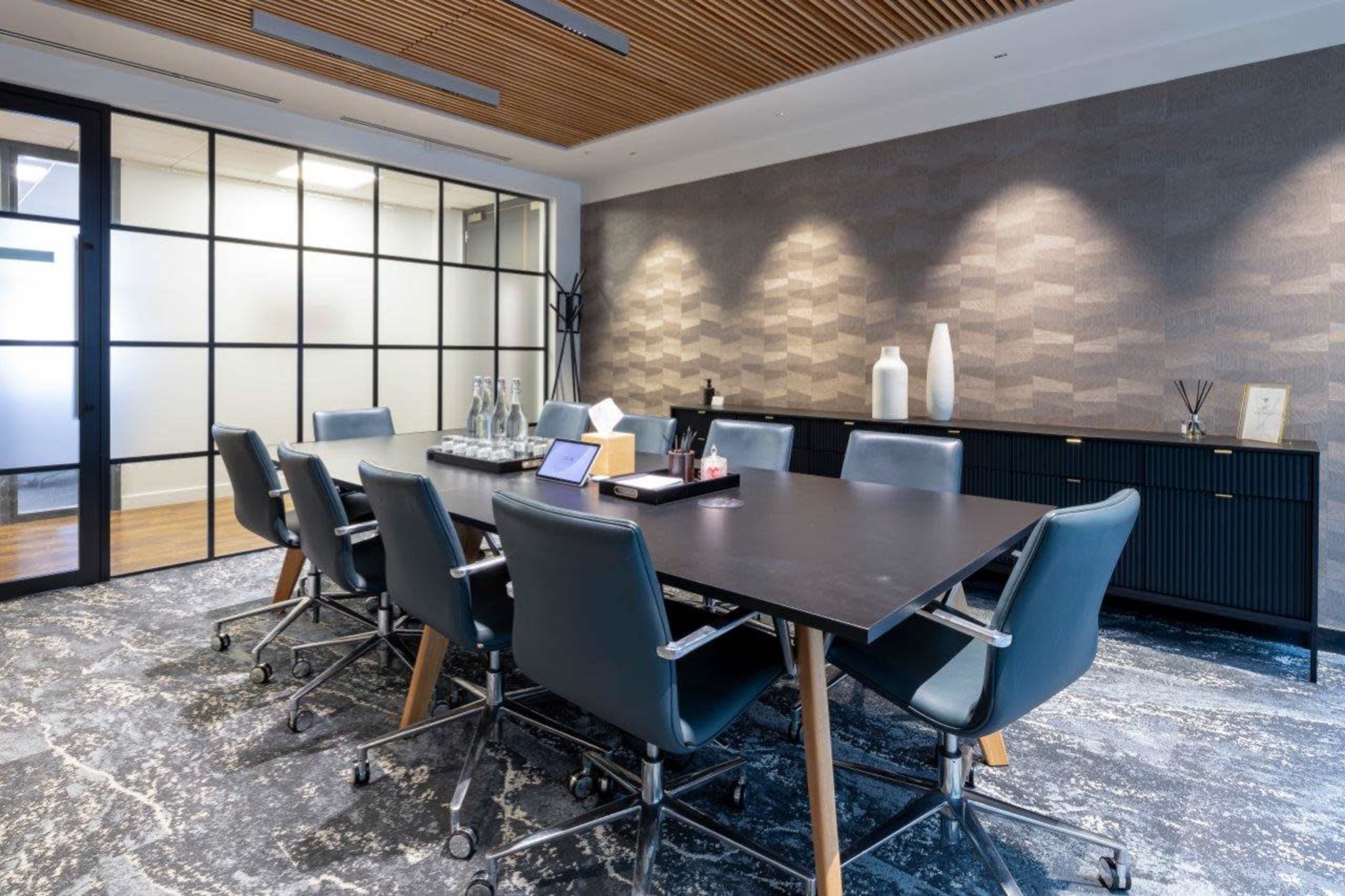 A modern conference room features a large rectangular table surrounded by ergonomic chairs, with a wall of glass partitions and textured décor.