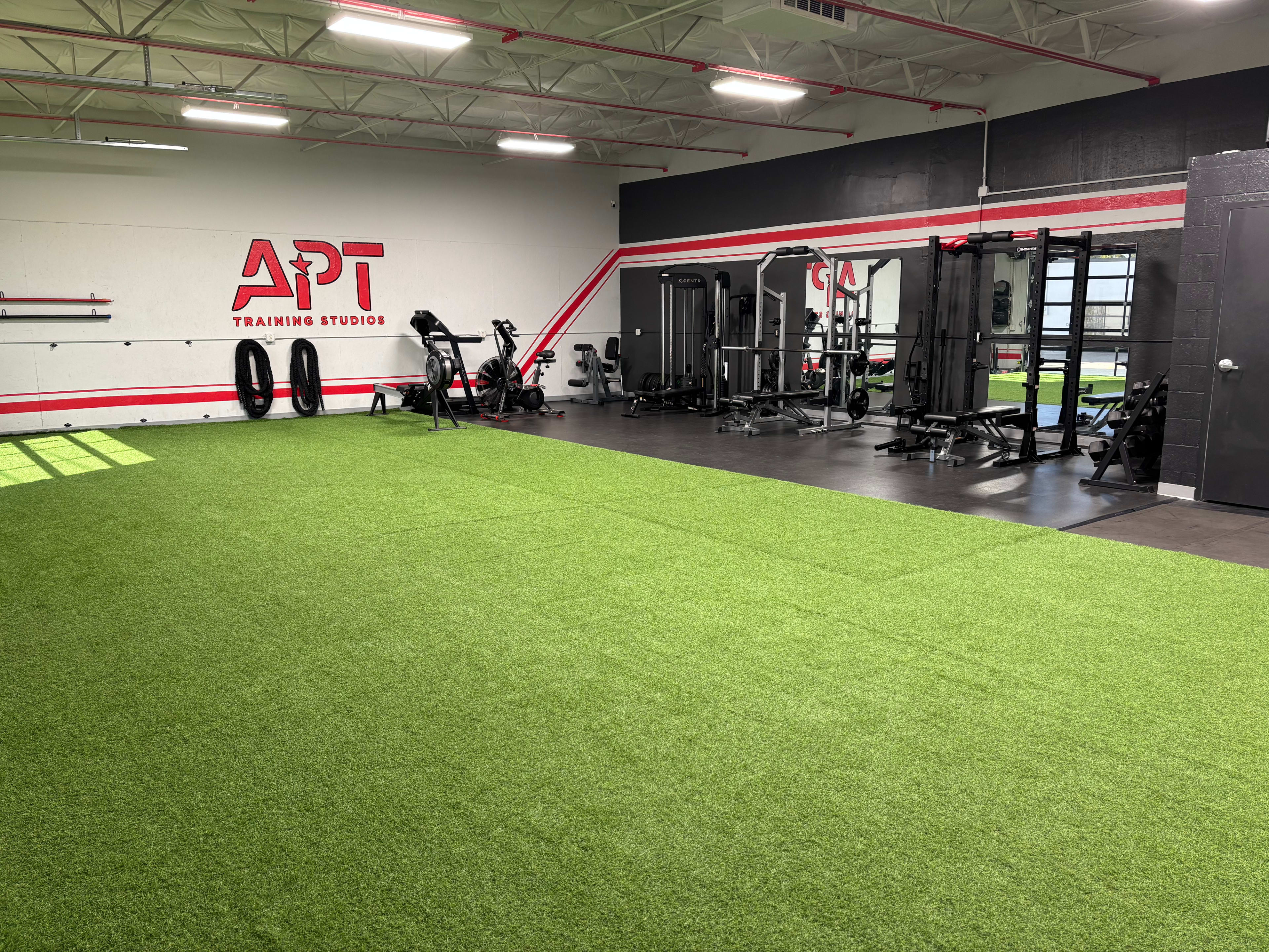 The image shows a gym space equipped with exercise machines, free weights, and a turf area for training.