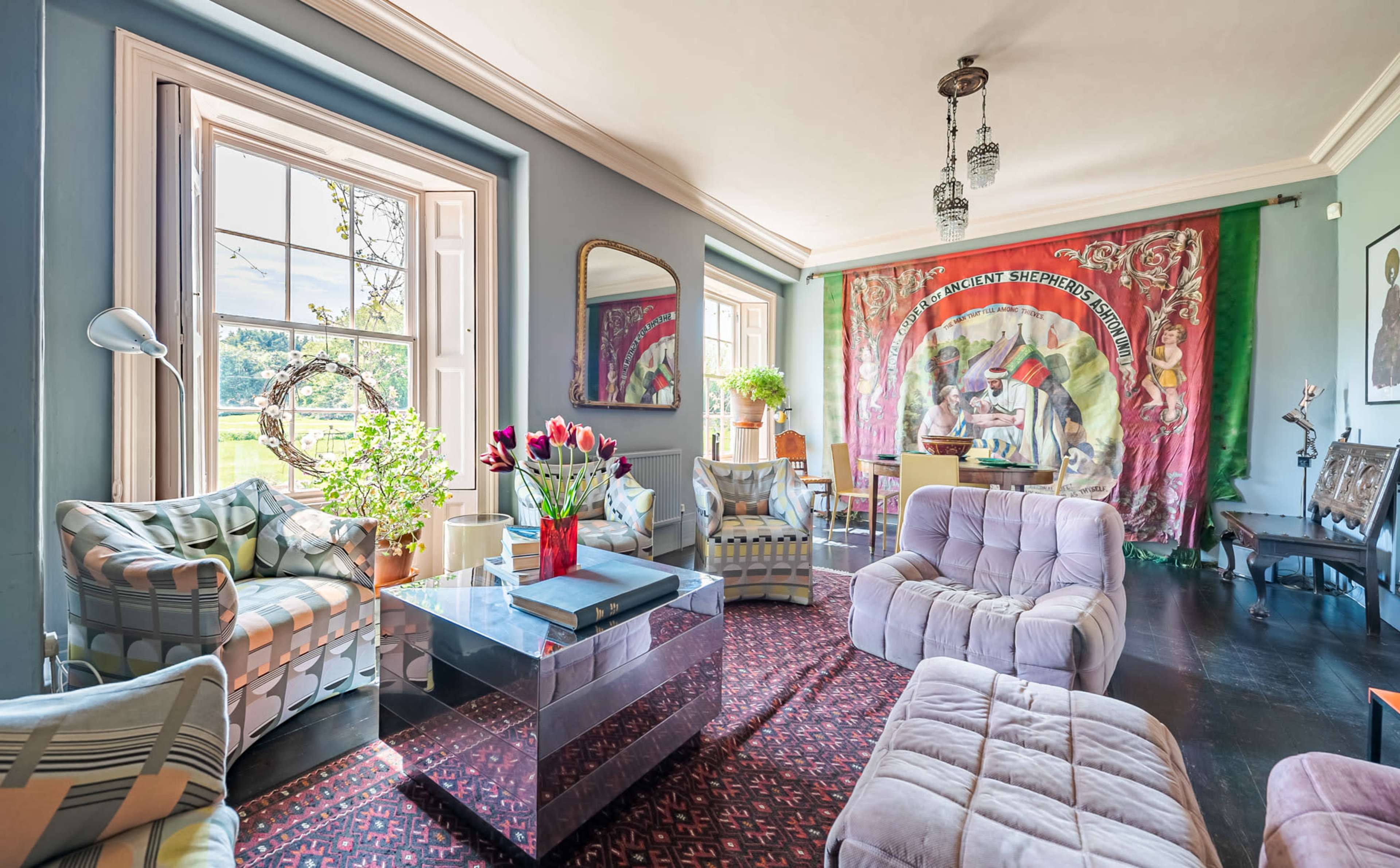 A brightly lit living room featuring modern furniture, a floral arrangement on a glass table, and a large colorful wall tapestry.