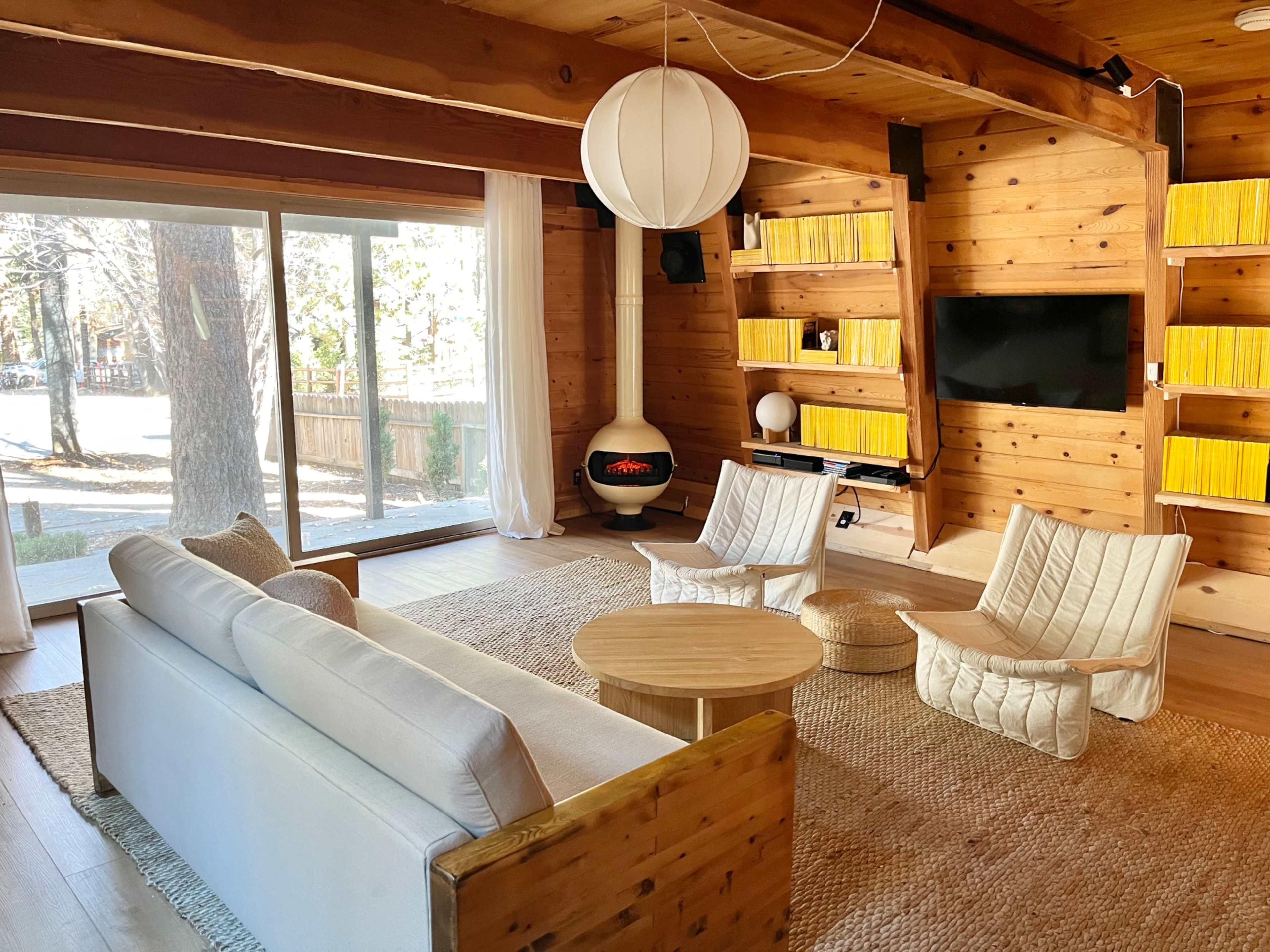 The image shows a modern wooden living room with a round coffee table, two armchairs, a sofa, and a fireplace, all arranged near large windows that let in natural light.