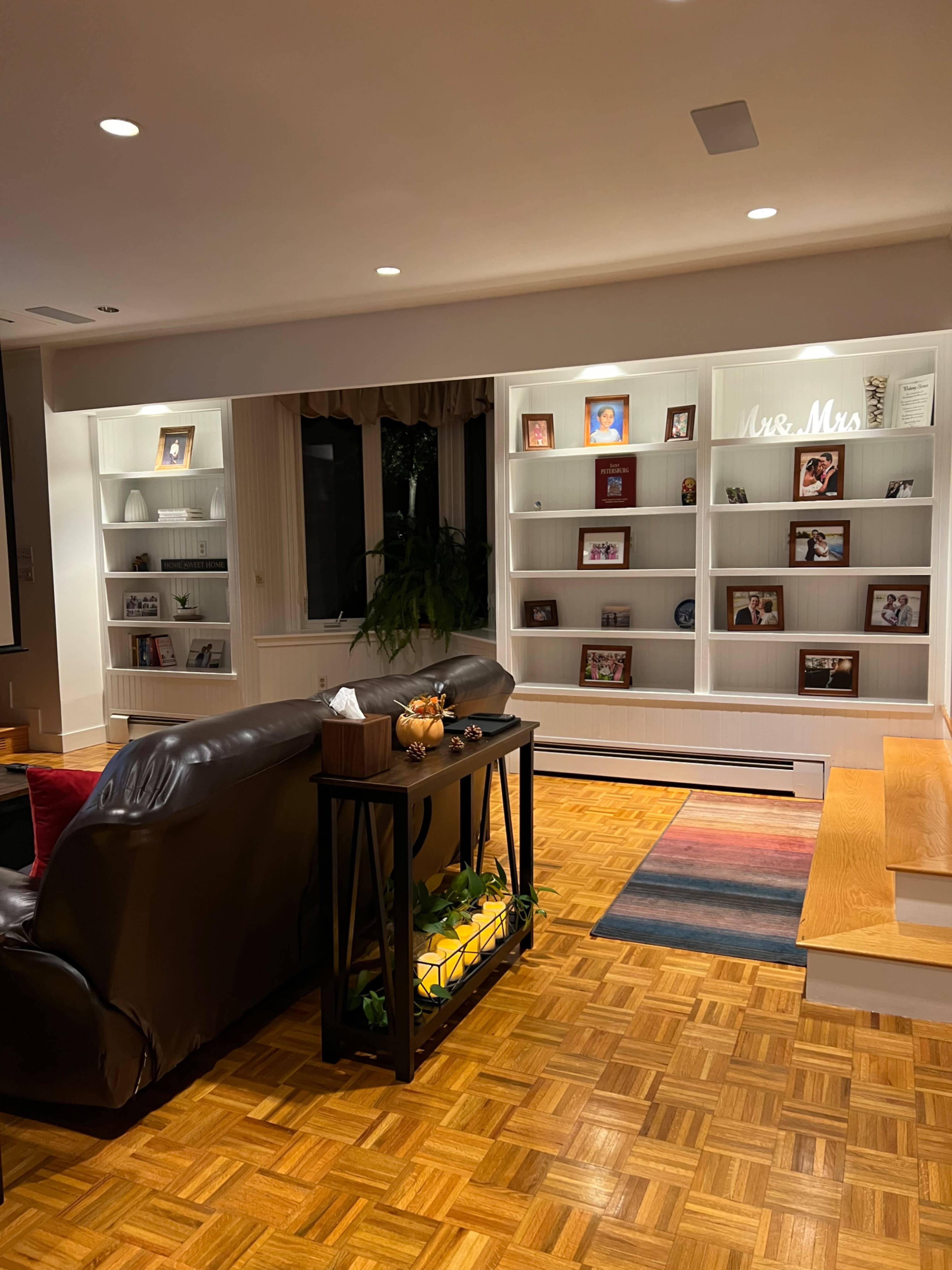 A living room features a dark leather sofa facing a white shelving unit with framed photos and decorations, set against a patterned wood floor.
