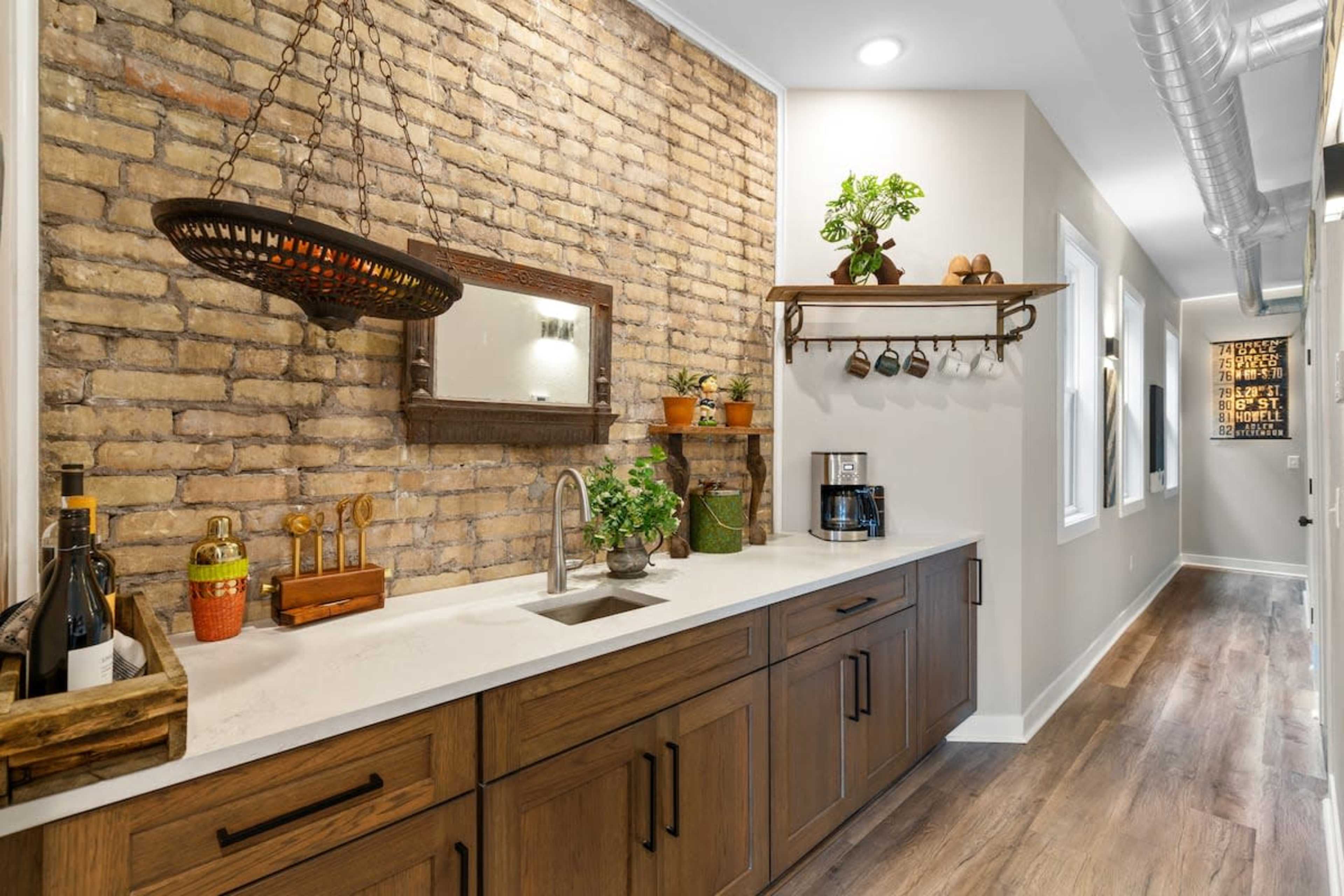 The image shows a hallway with a brick accent wall, a wooden countertop featuring a sink, and various plants and kitchen items arranged on shelves above.