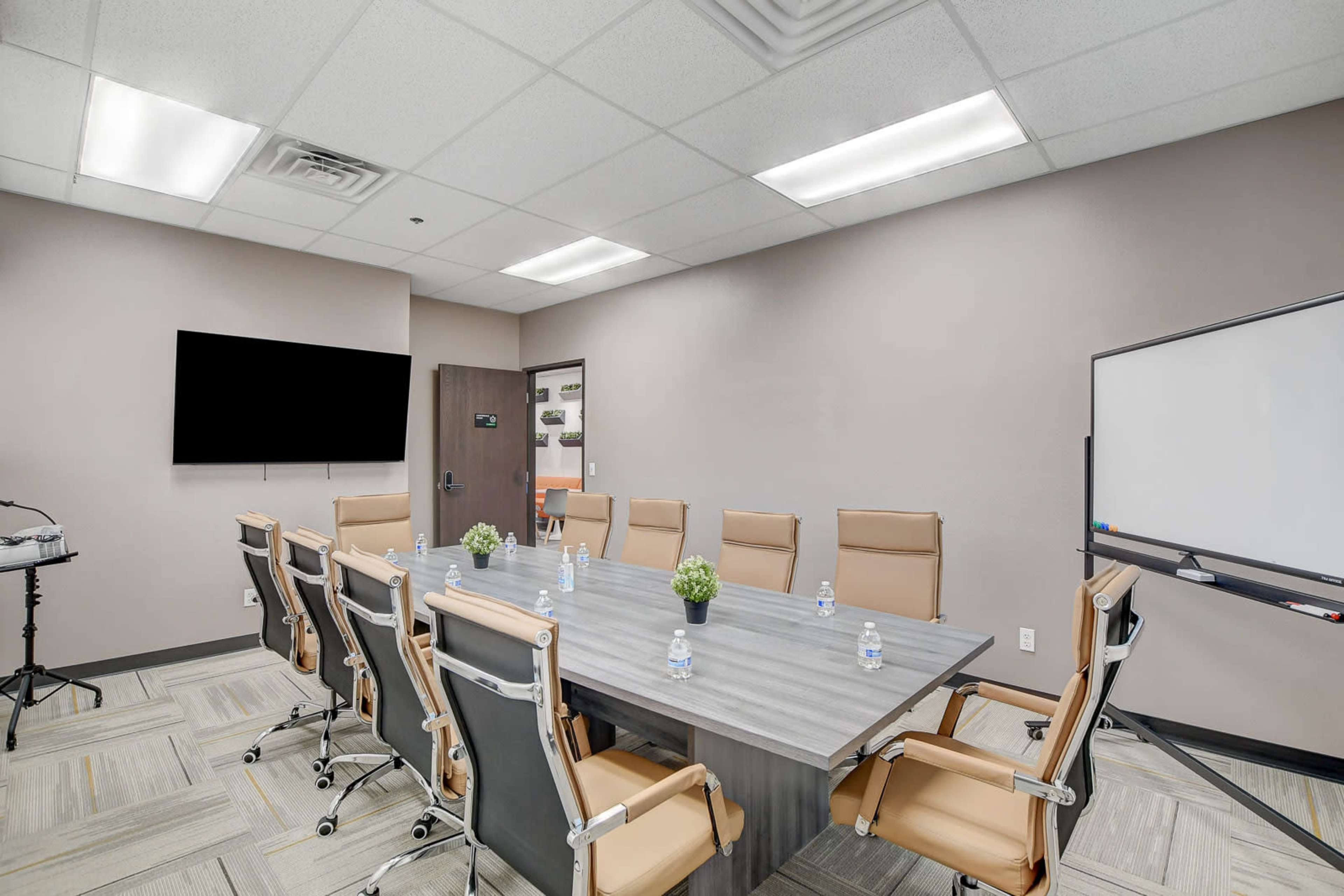A modern conference room features a large table surrounded by ergonomic chairs, with a television screen, whiteboard, and small decorative plants.