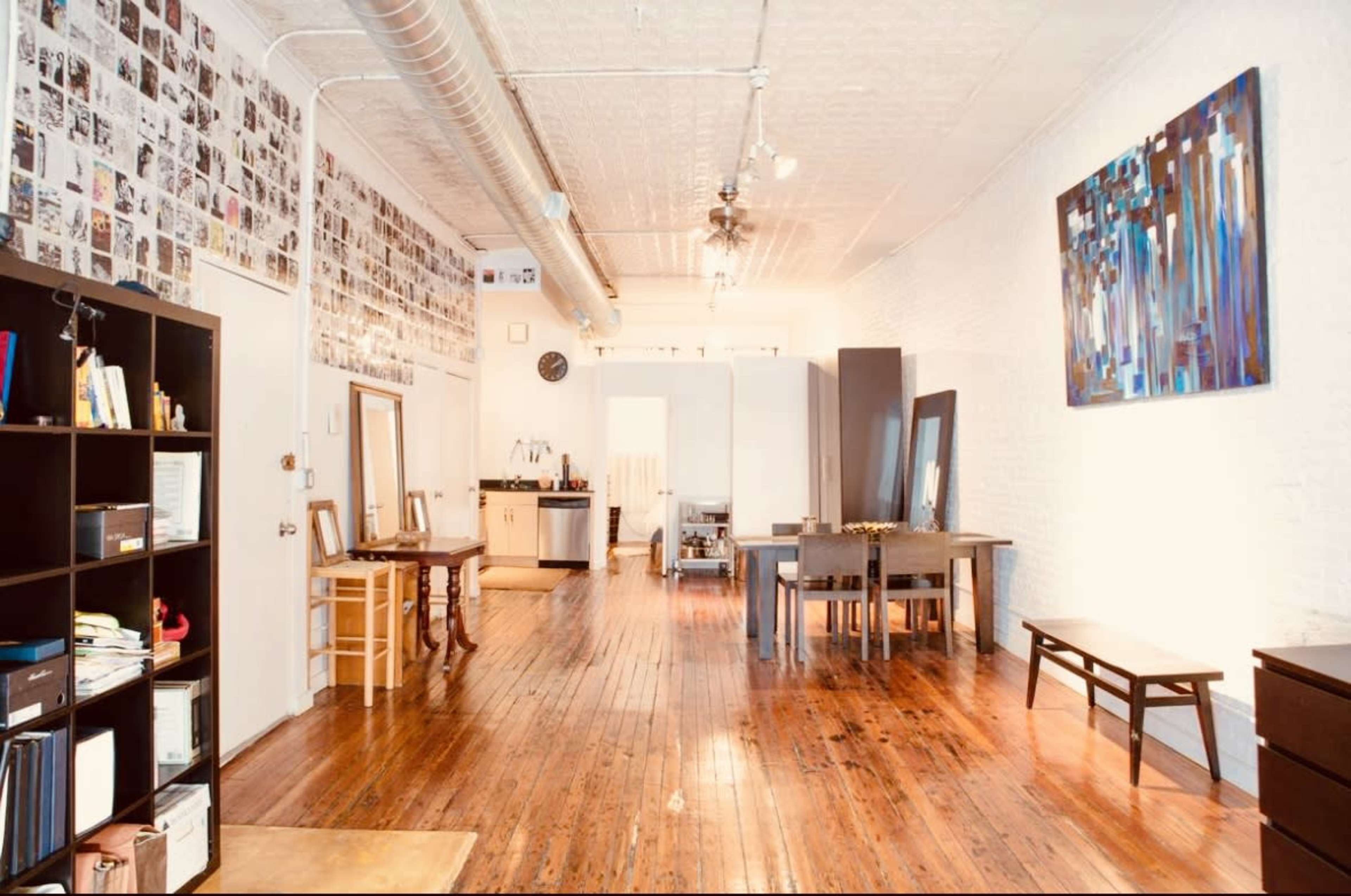 The image shows a spacious interior of an apartment with wooden flooring, white brick walls adorned with black and white photographs, and furniture arranged in a living area that includes a dining table and two mirrors.