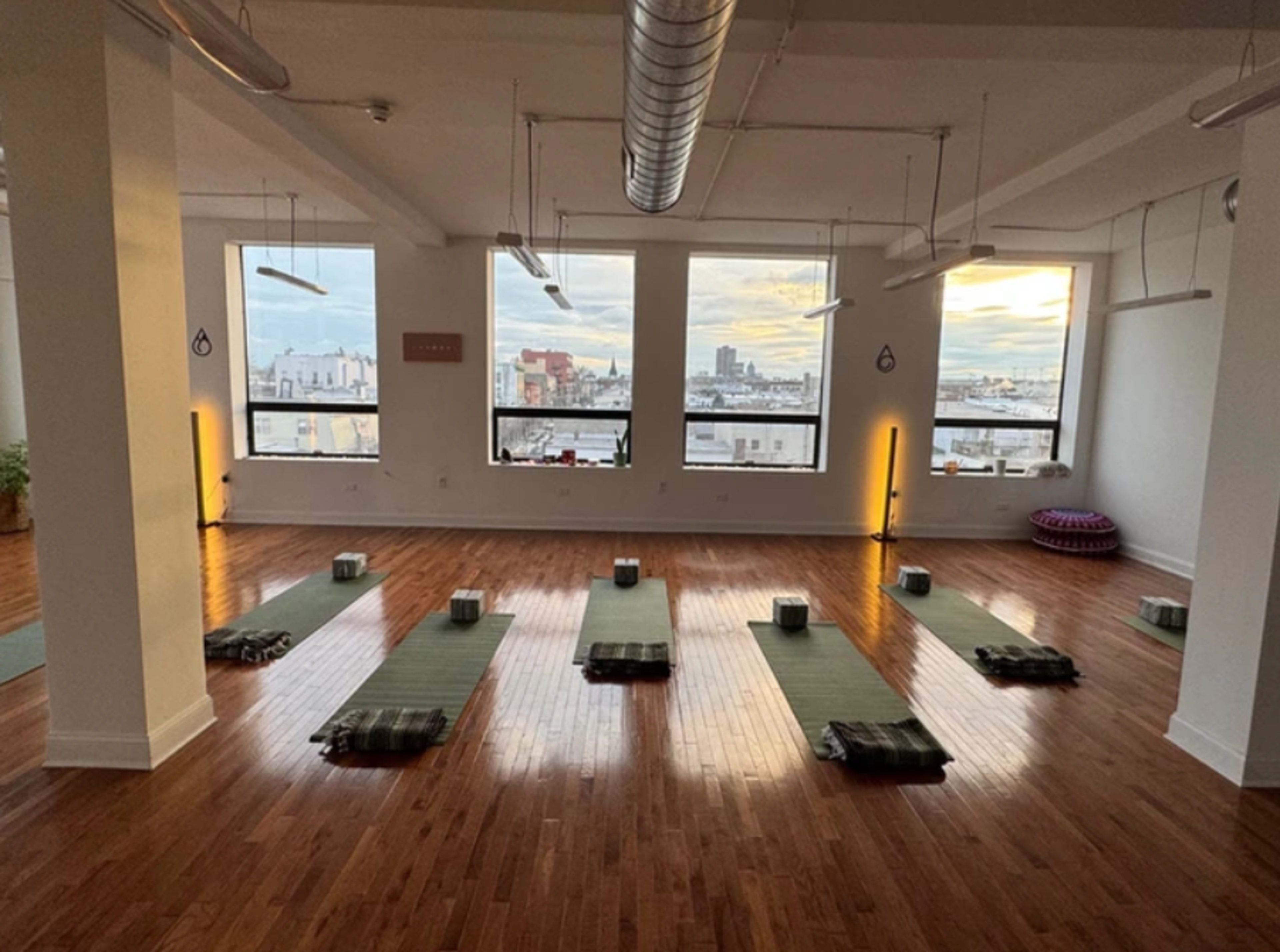 The image shows a spacious yoga studio with green mats arranged neatly on a polished wooden floor and large windows displaying an urban skyline.