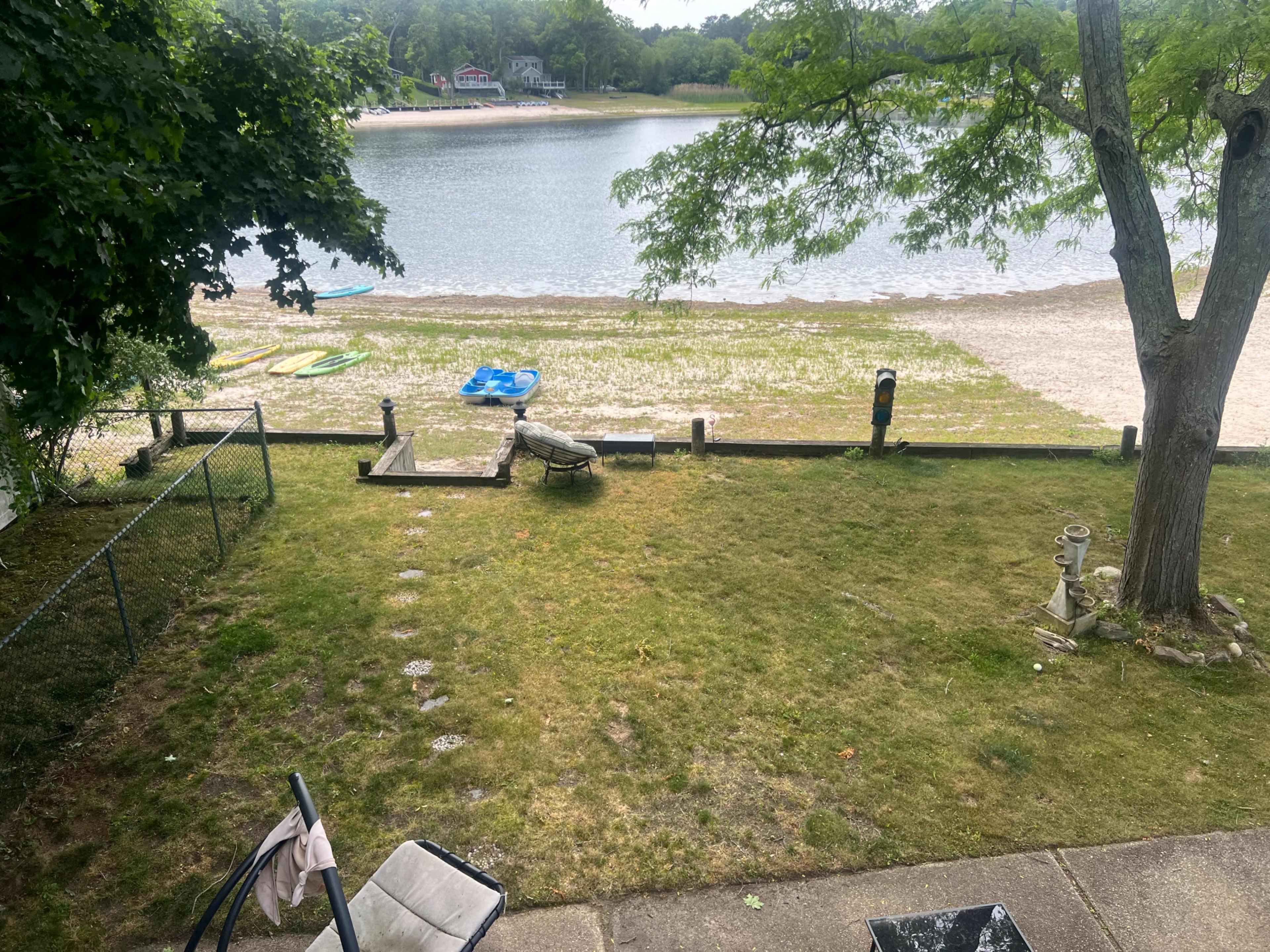 The image shows a grassy area leading to a sandy beach along a calm lake, with kayaks and a few trees in the foreground.