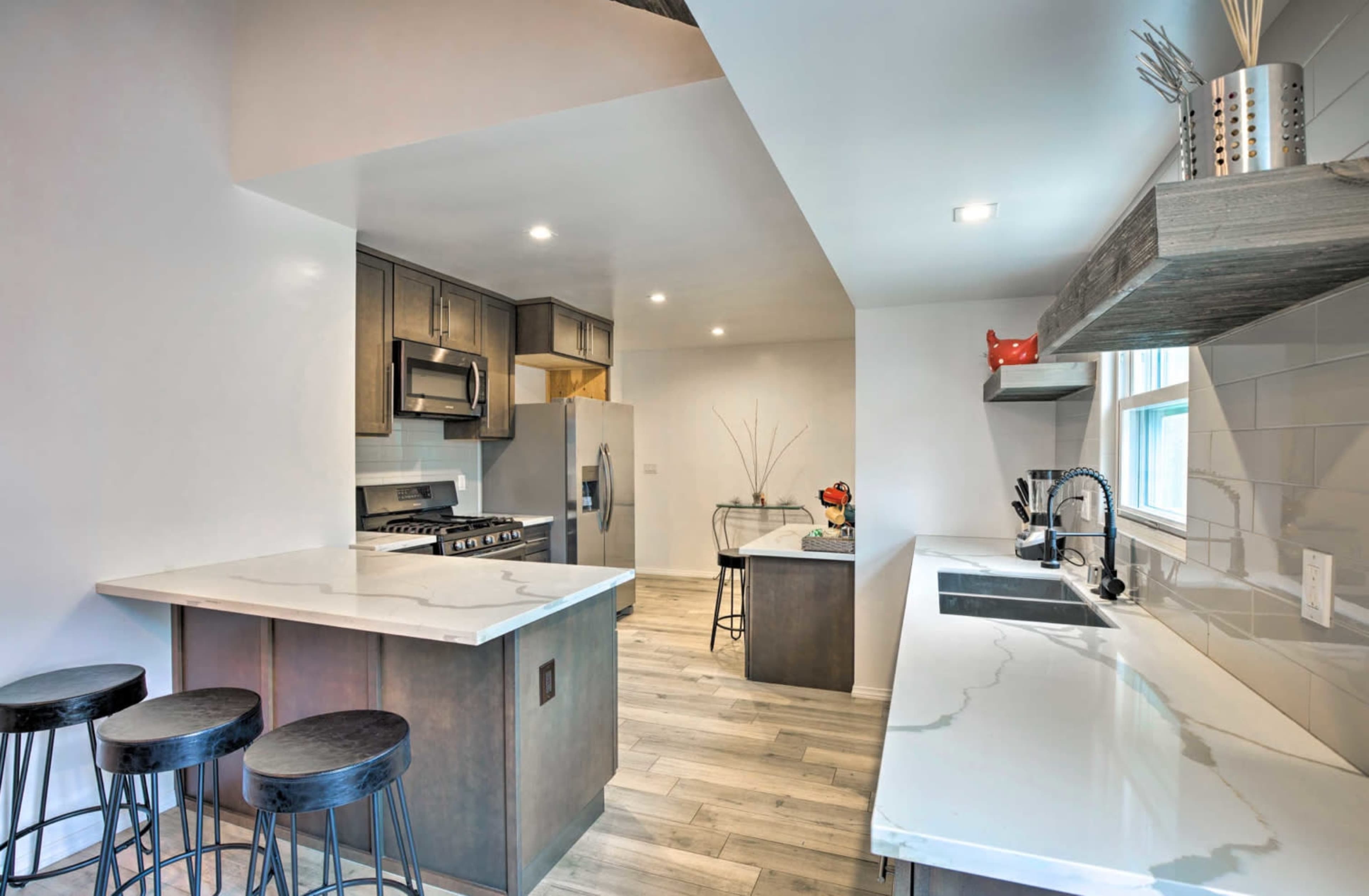The image shows a modern kitchen featuring a white countertop with barstools, stainless steel appliances, and wooden flooring.