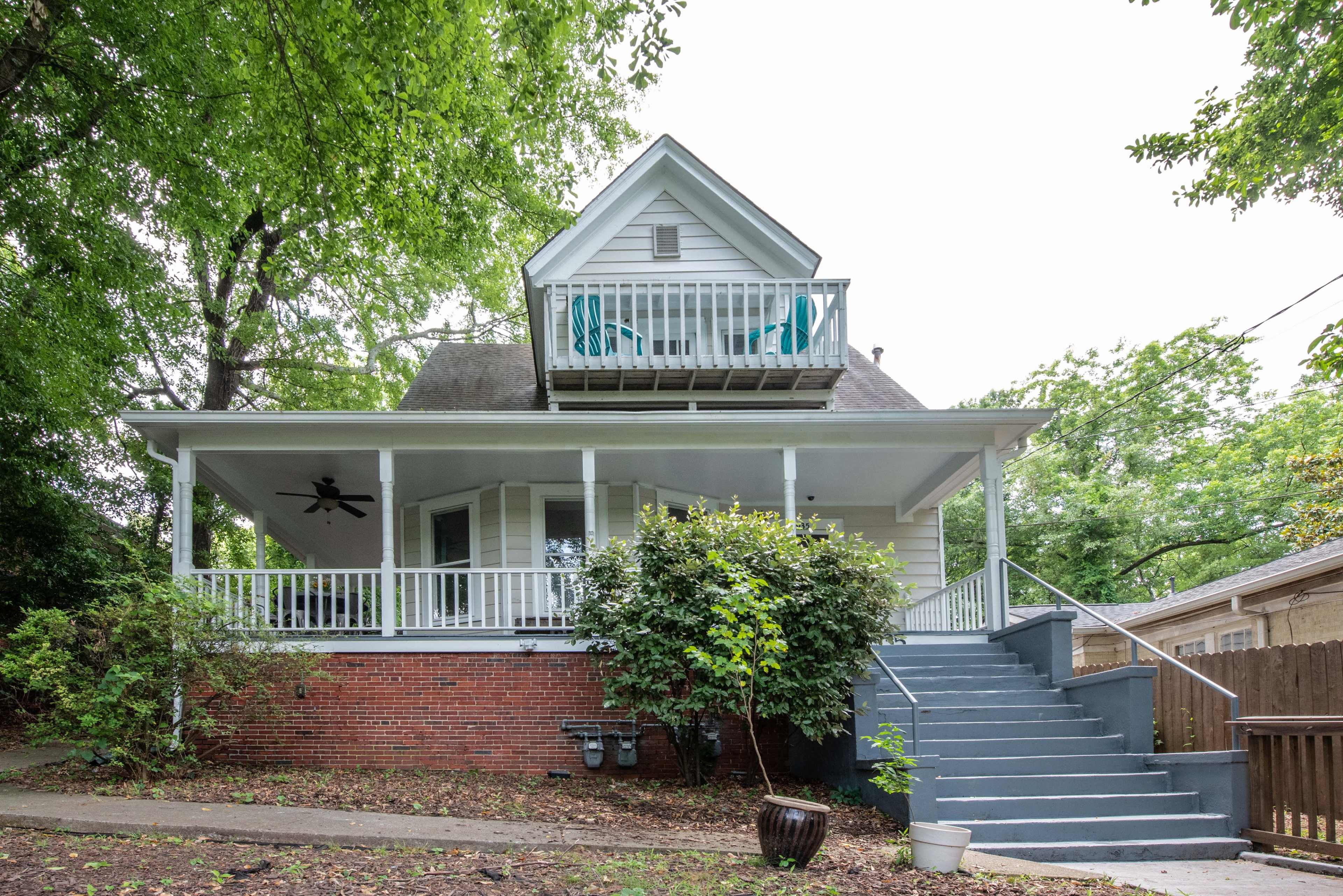 A two-story white house with a covered porch and a balcony, surrounded by trees and a pathway leading to the entrance.