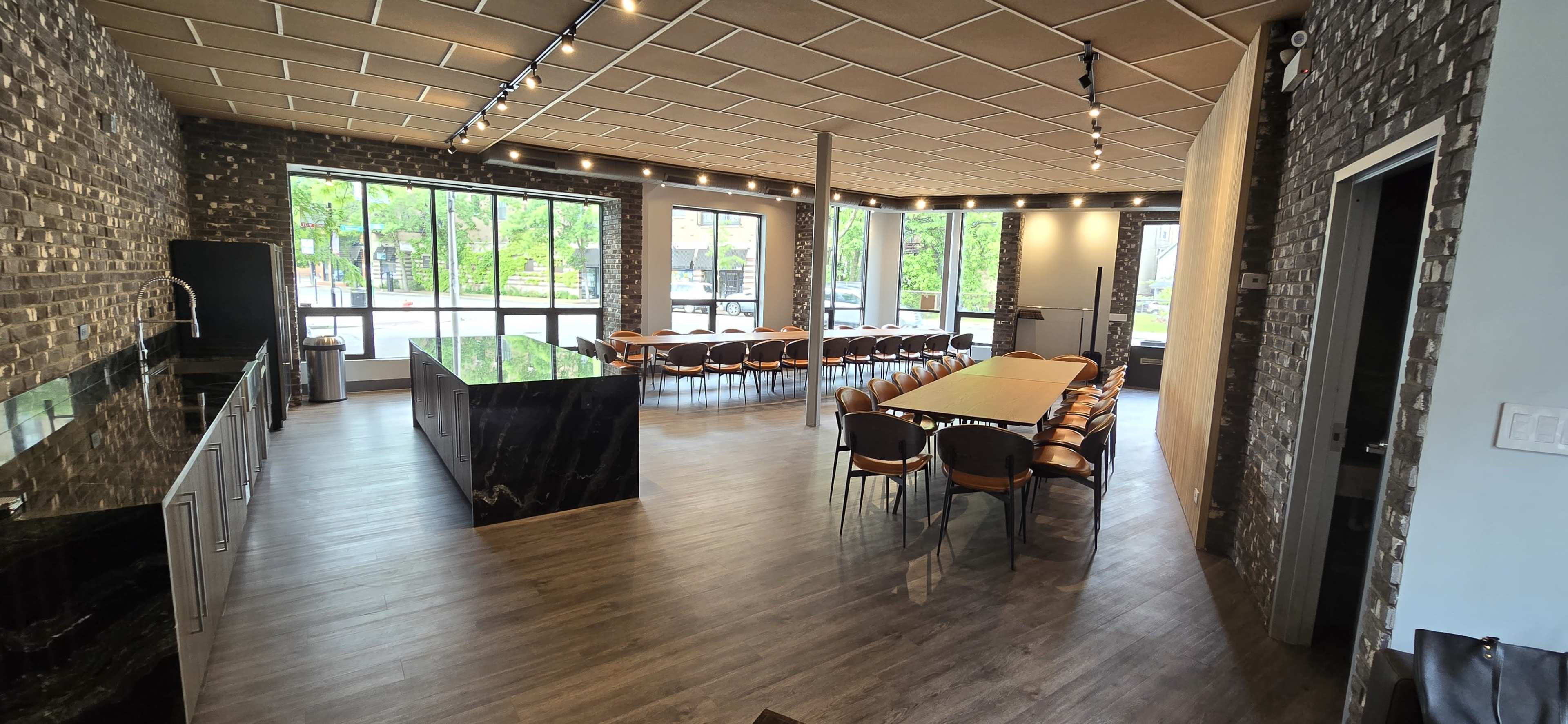 A modern interior space featuring a large dining area with a long table and rows of chairs, surrounded by large windows and stone walls.