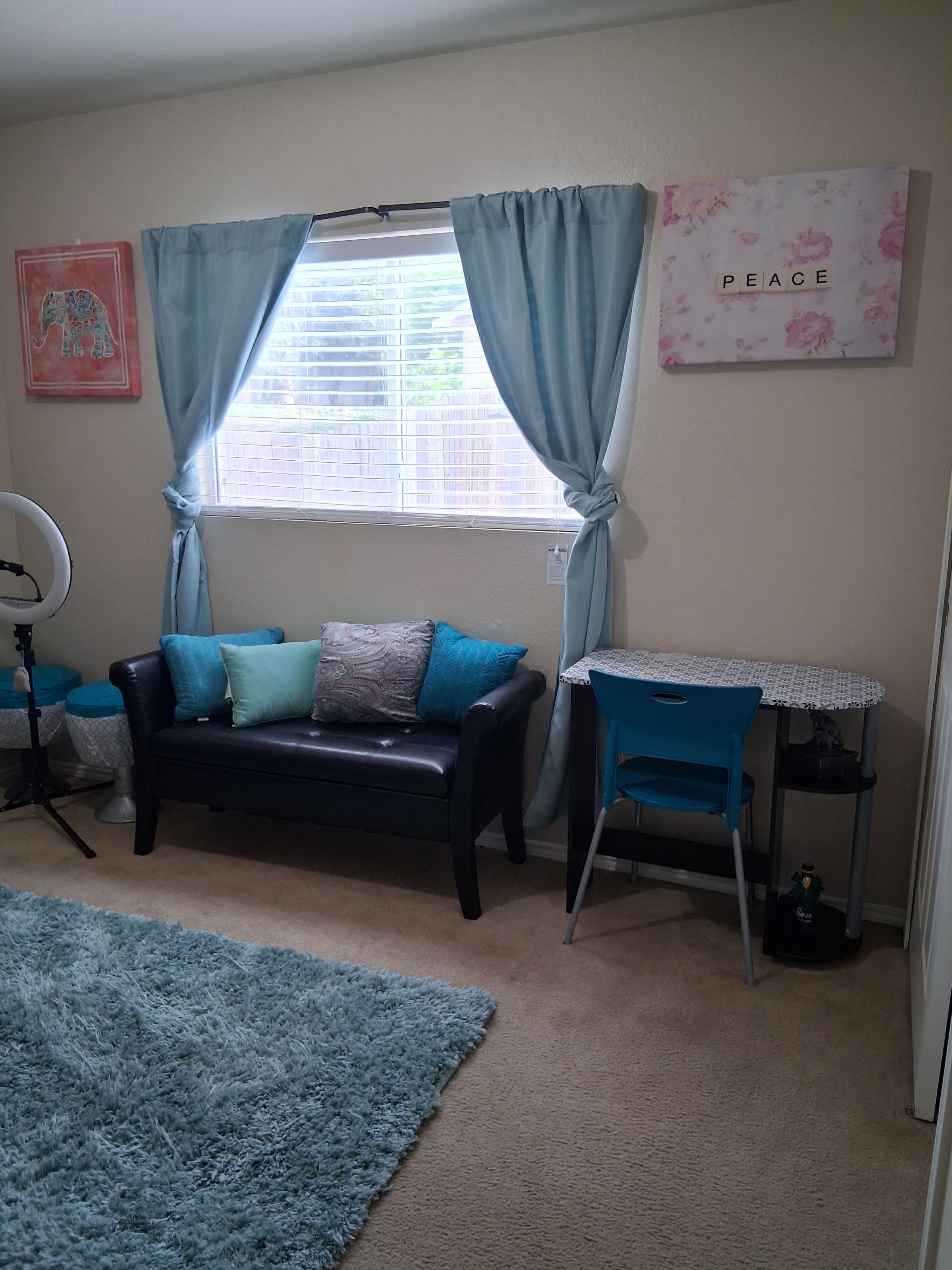 The image shows a cozy room with a black sofa adorned with teal and white pillows, a small desk with a blue chair, and light blue curtains framing a window.