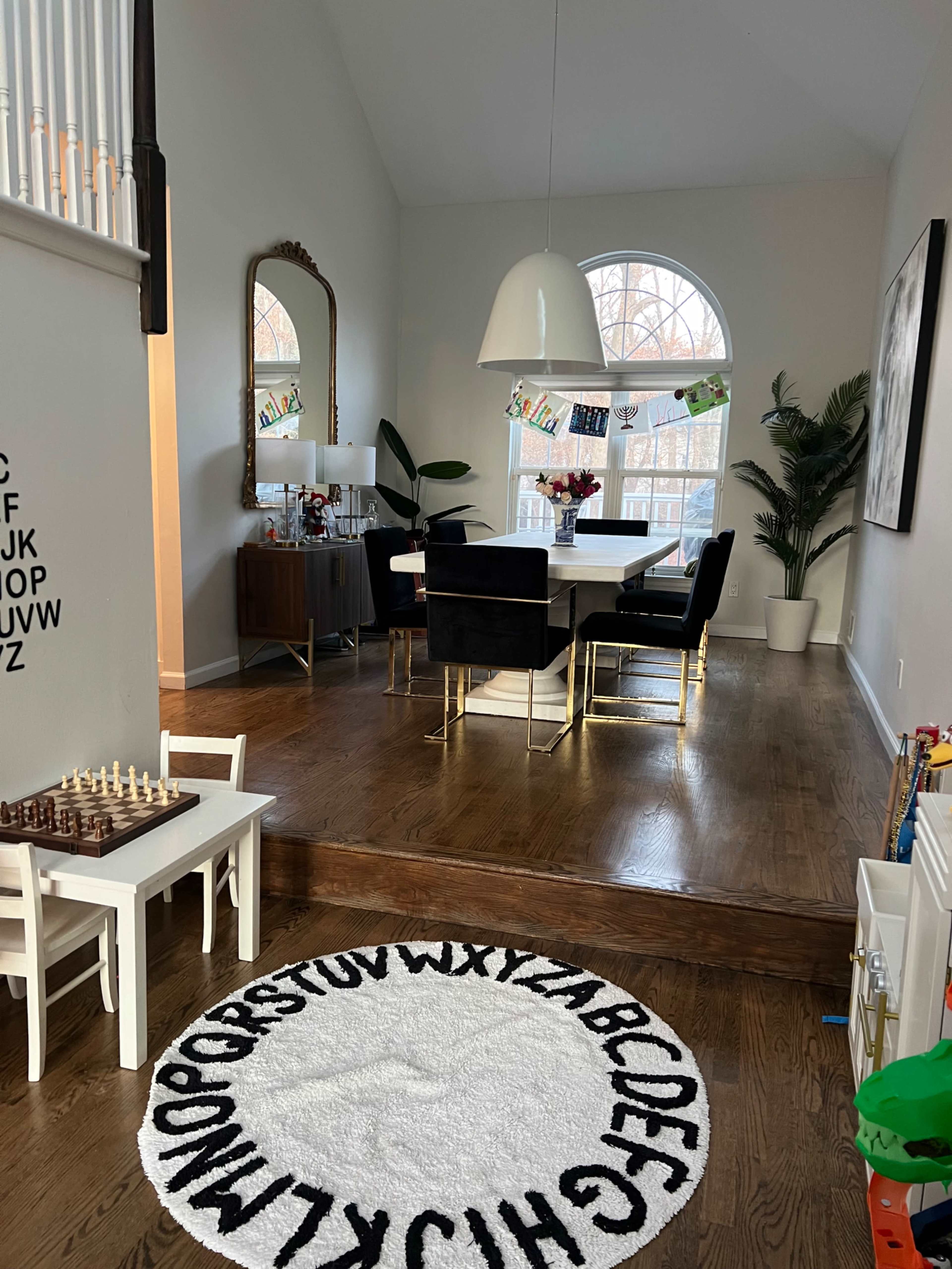 A dining area features a modern table surrounded by black chairs, with a large light fixture overhead and a decorative mirror on the wall.