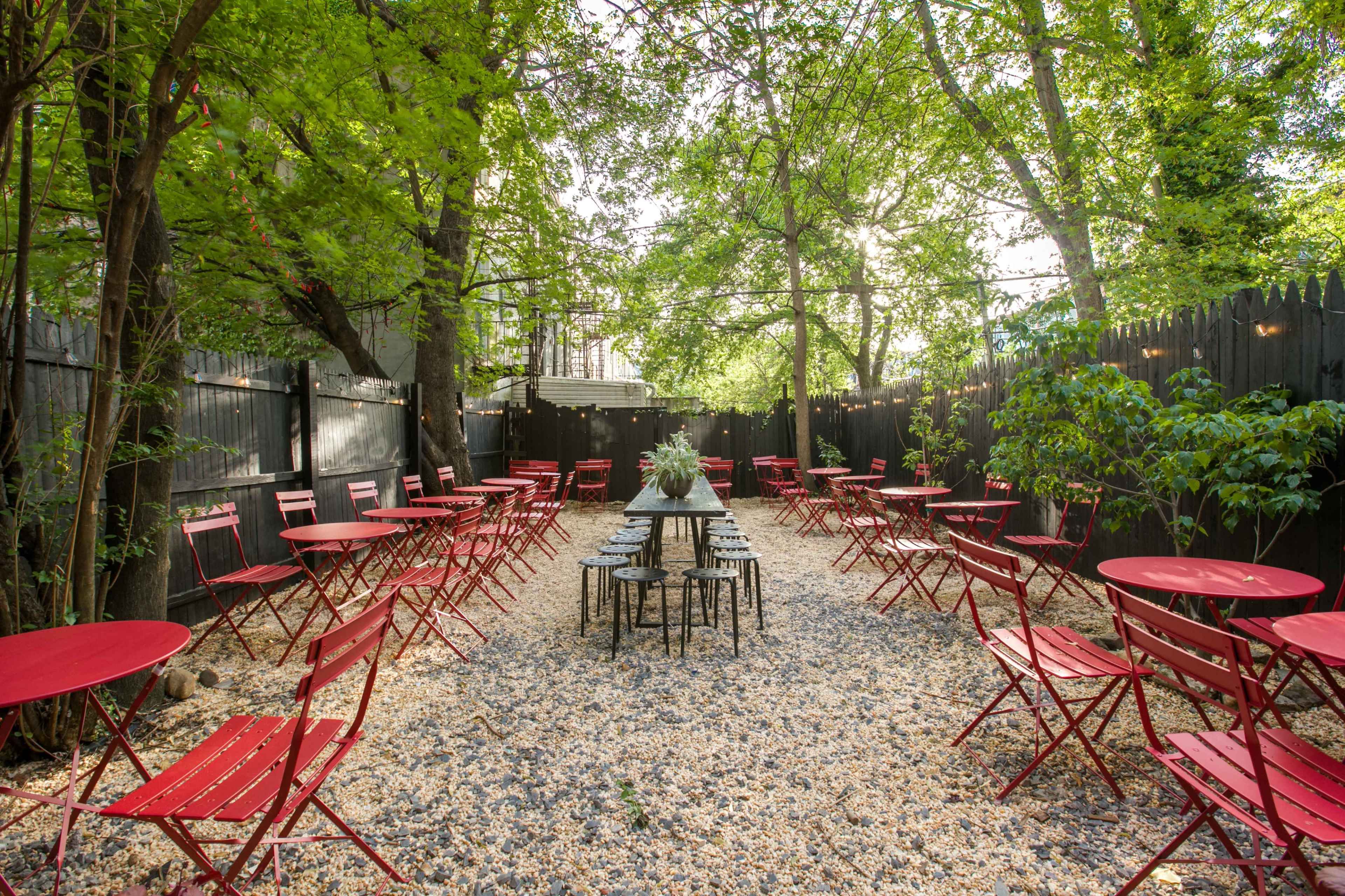 A garden café features red metal tables and chairs arranged on a gravel path surrounded by trees.