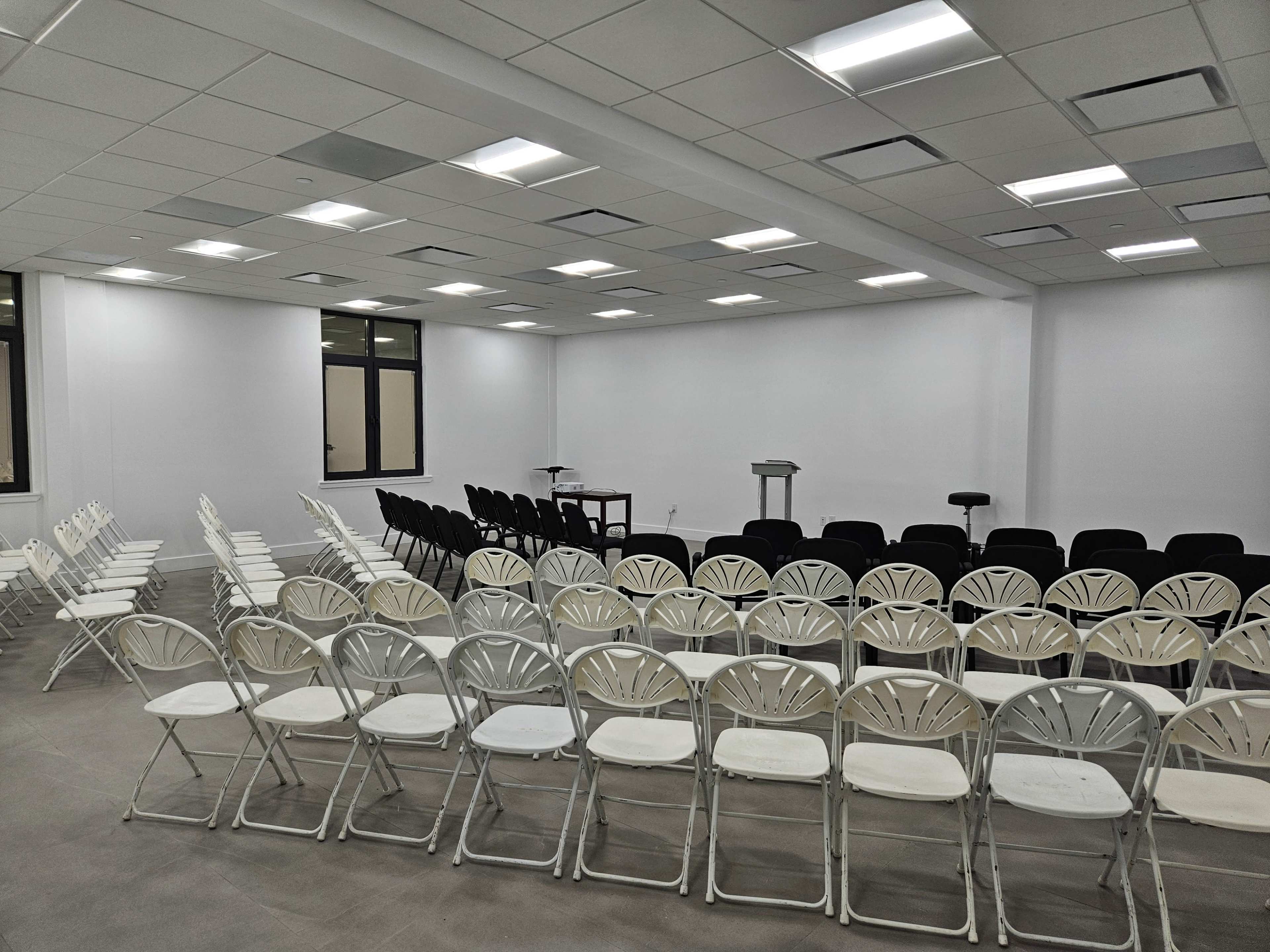 A large meeting room is set up with rows of white and black folding chairs facing a podium and a small table.