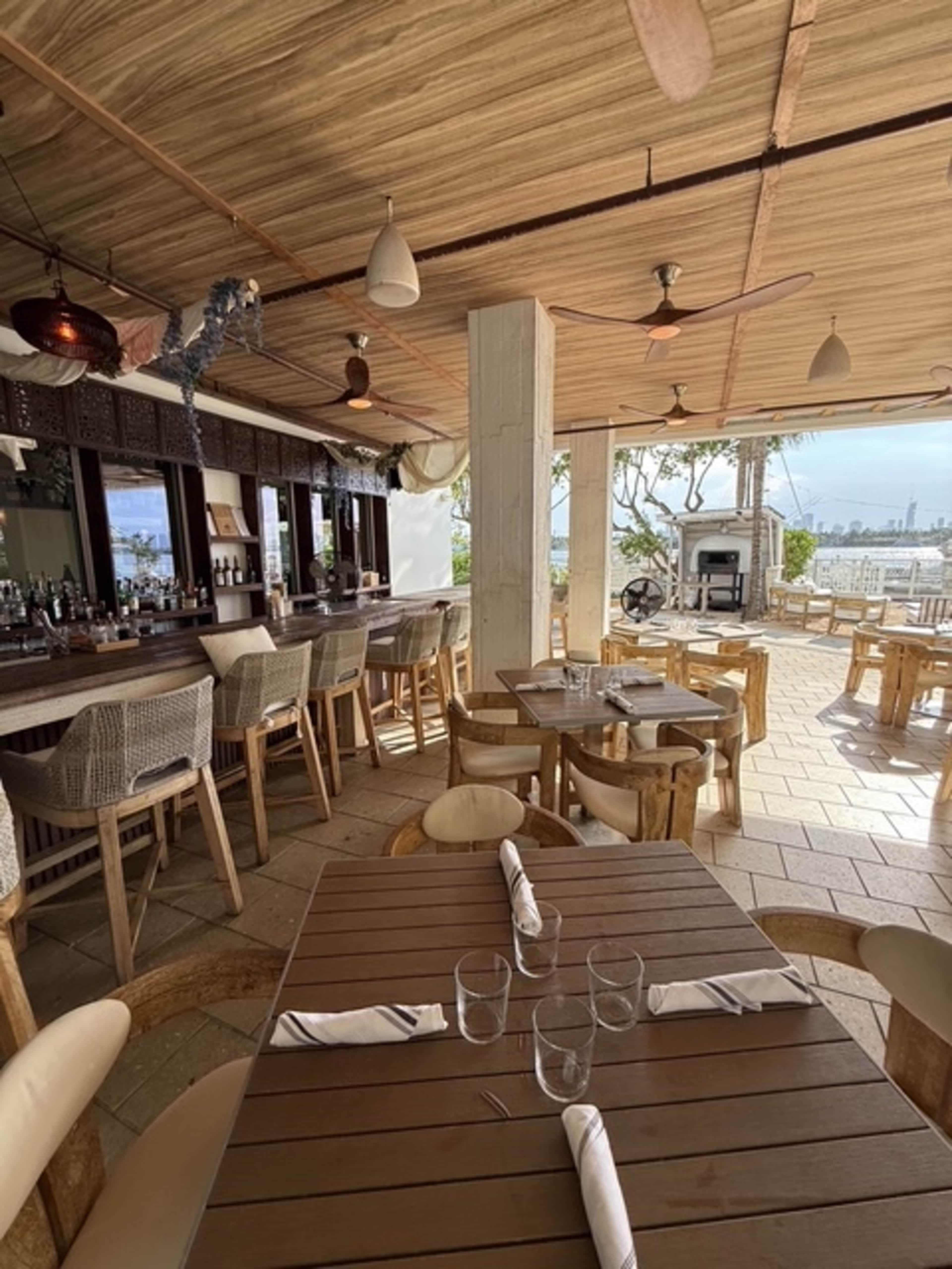 The image shows a partially outdoor restaurant with wooden tables and chairs, a bar area, and large windows overlooking a waterfront view.