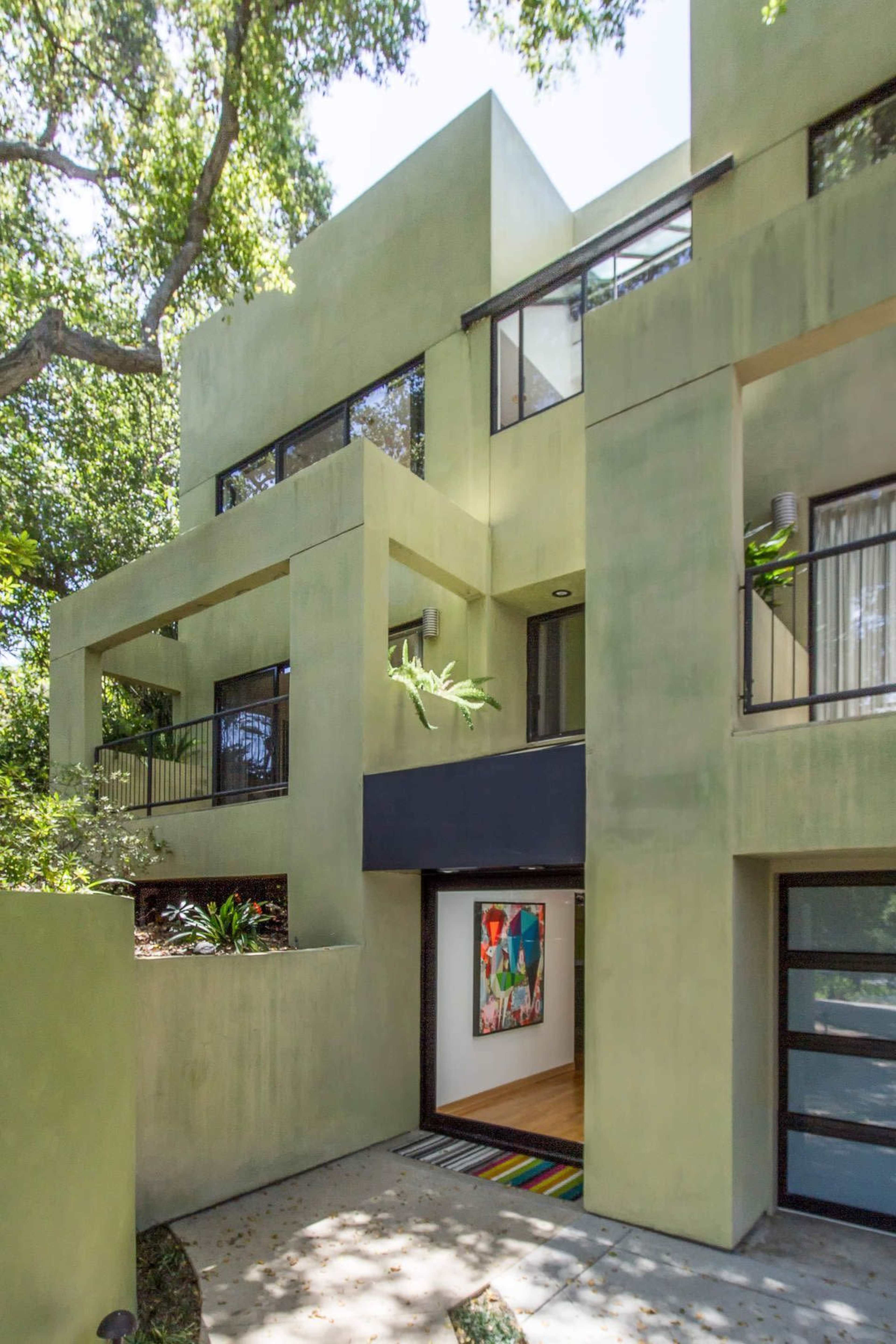 The image shows a modern, multi-level house with a green exterior and large windows surrounded by trees.