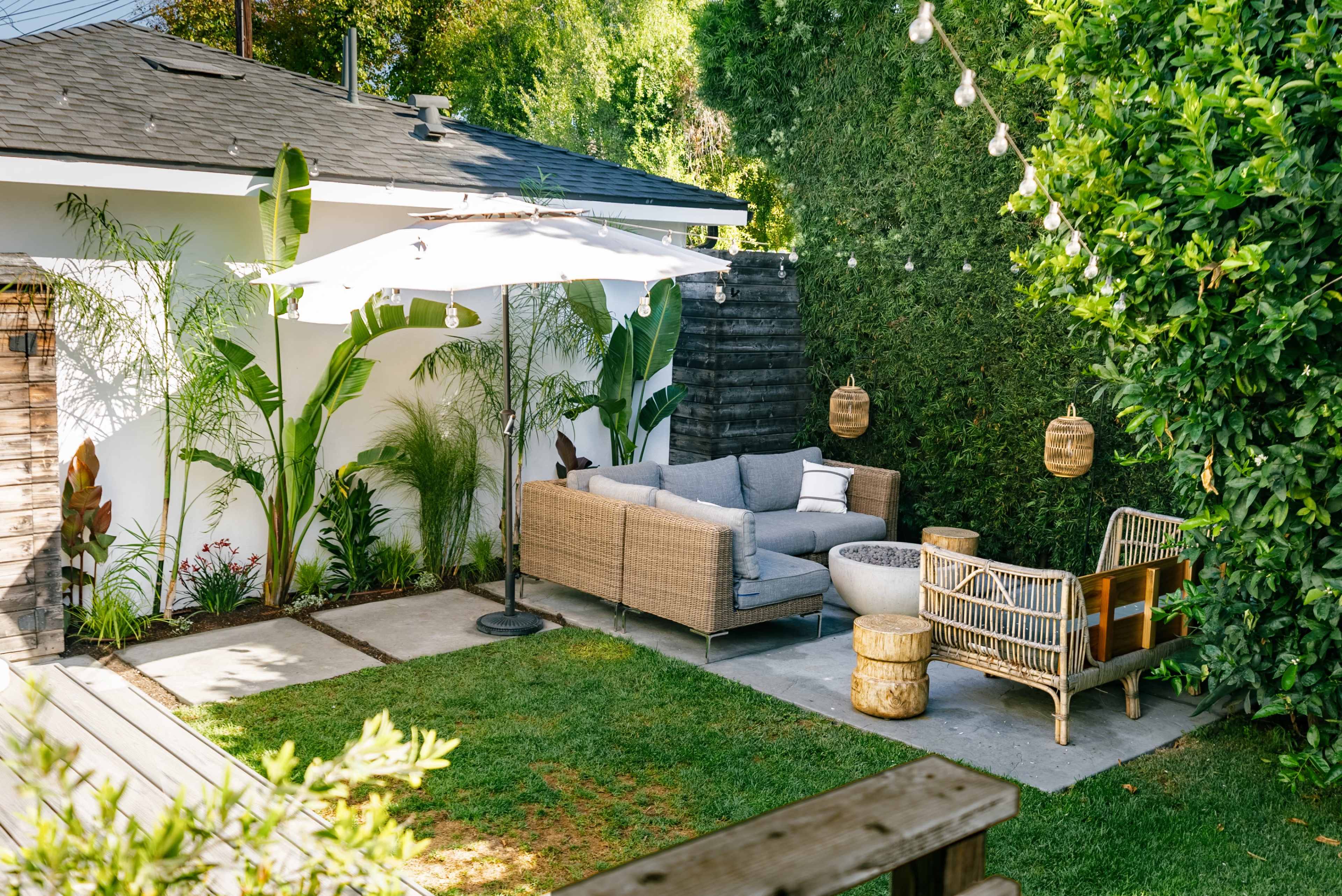 A patio area with a wicker sofa, a sun umbrella, a small table, and two decorative chairs surrounded by greenery.