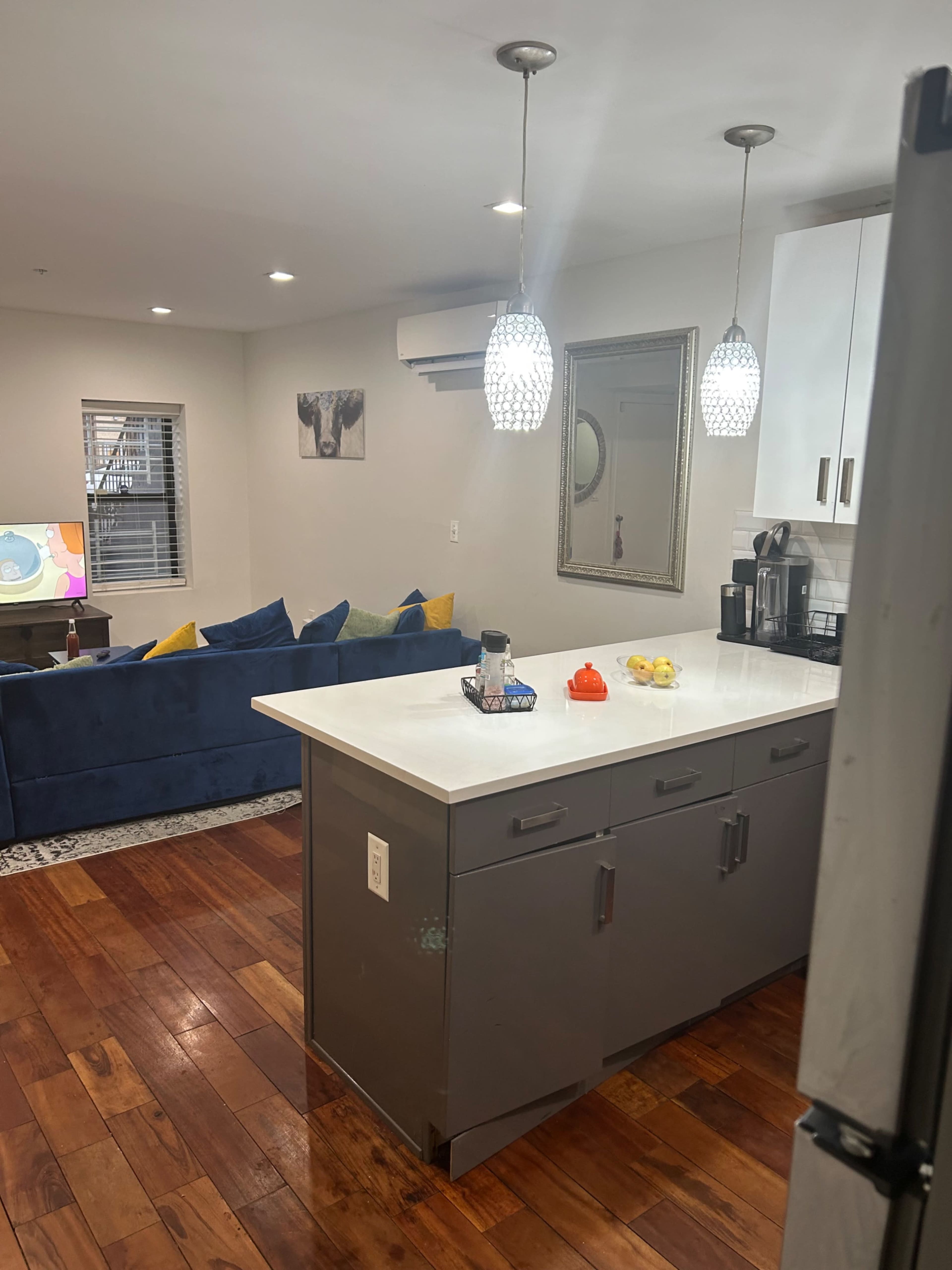 The image shows a modern kitchen area with a central island, gray cabinetry, a white countertop, and a living space in the background featuring a blue sofa and a television.