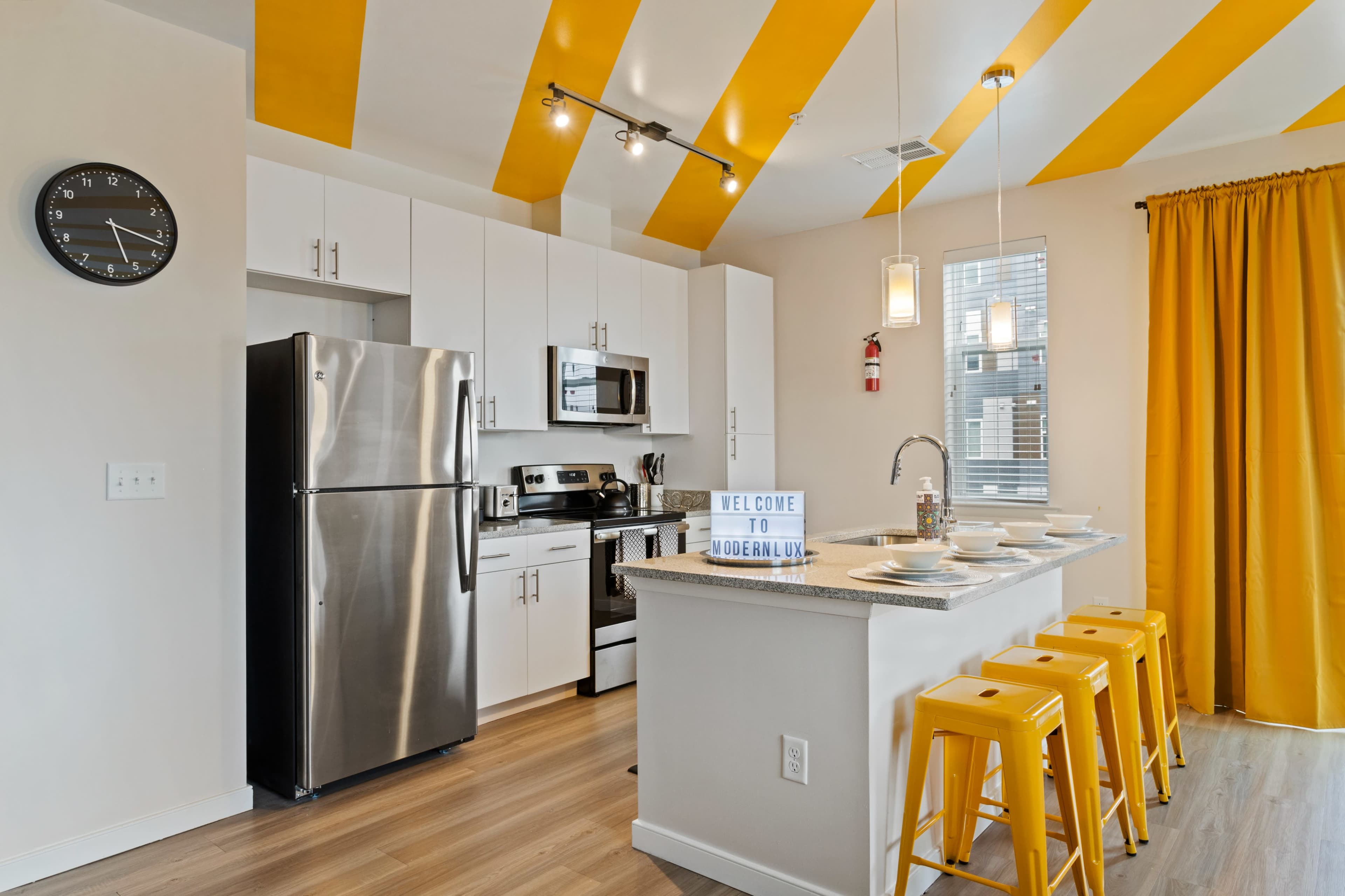 A modern kitchen features stainless steel appliances, yellow accents, and a breakfast bar with yellow stools.