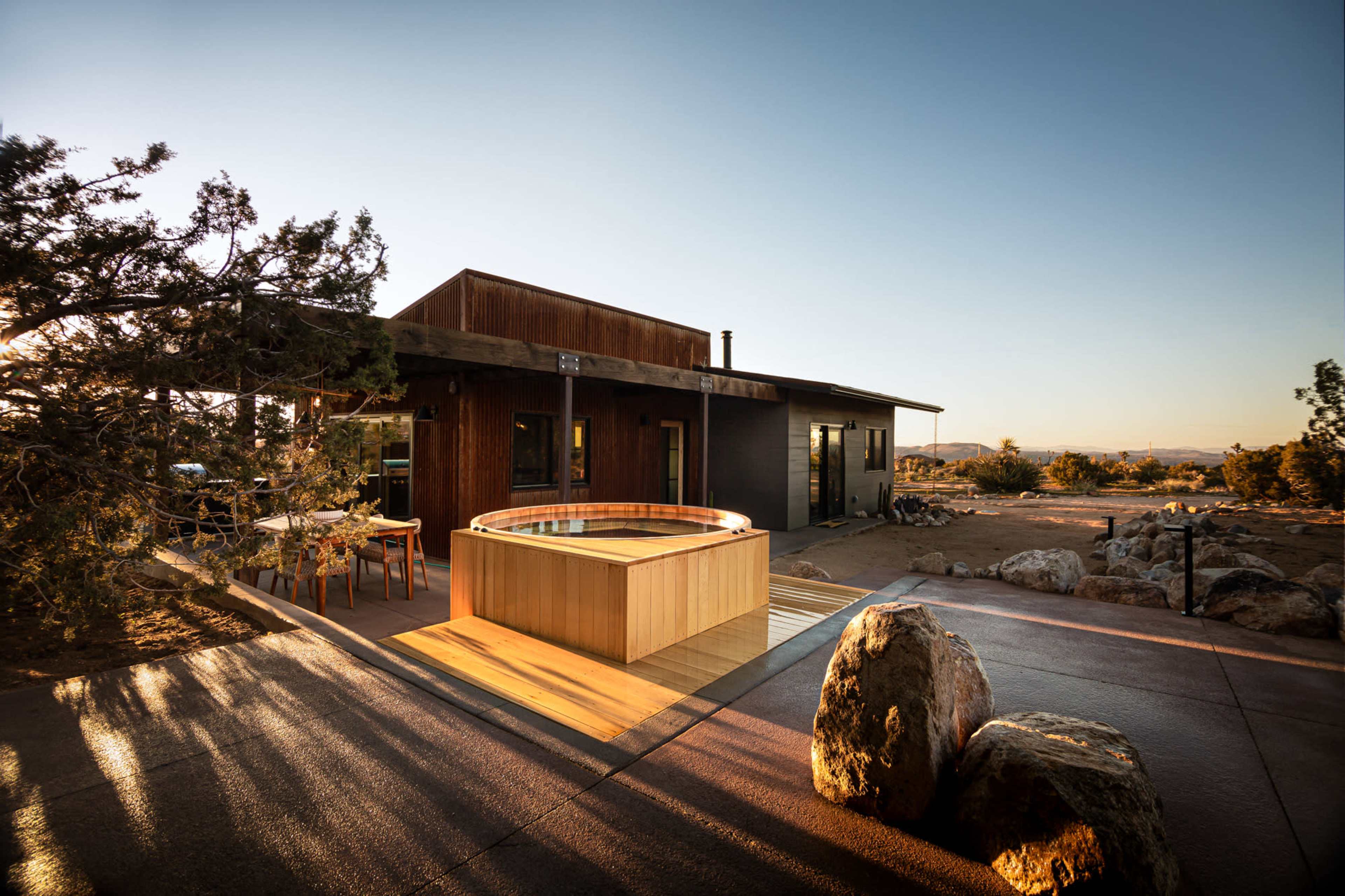 A modern cabin features a wooden hot tub on a patio surrounded by rocks and desert vegetation.