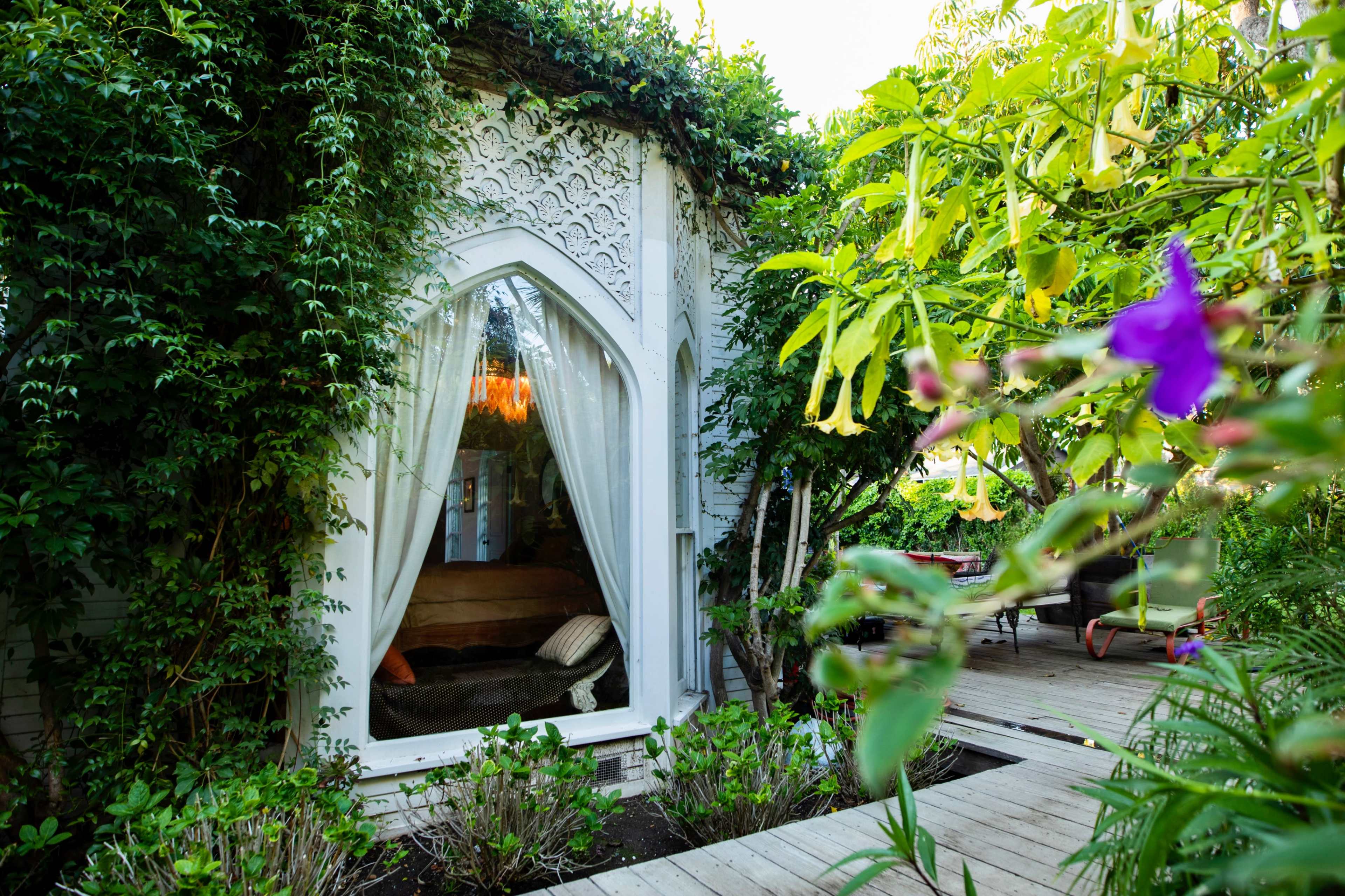 A decorative window framed by lush greenery opens to a cozy interior, surrounded by a wooden walkway and colorful plants.