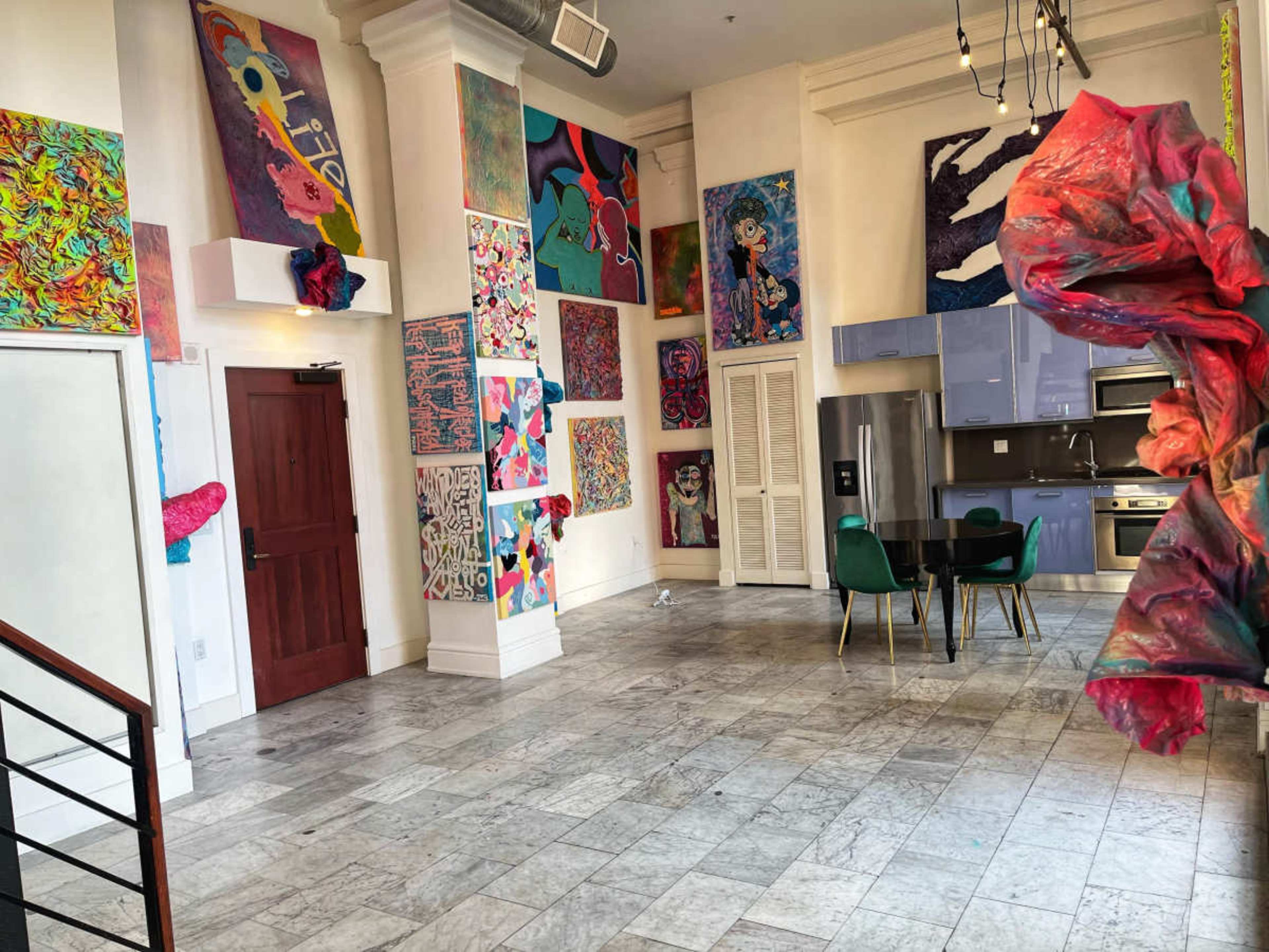 The image shows a spacious room with colorful abstract art pieces adorning the walls, a dining table with green chairs, and modern kitchen appliances in the background.