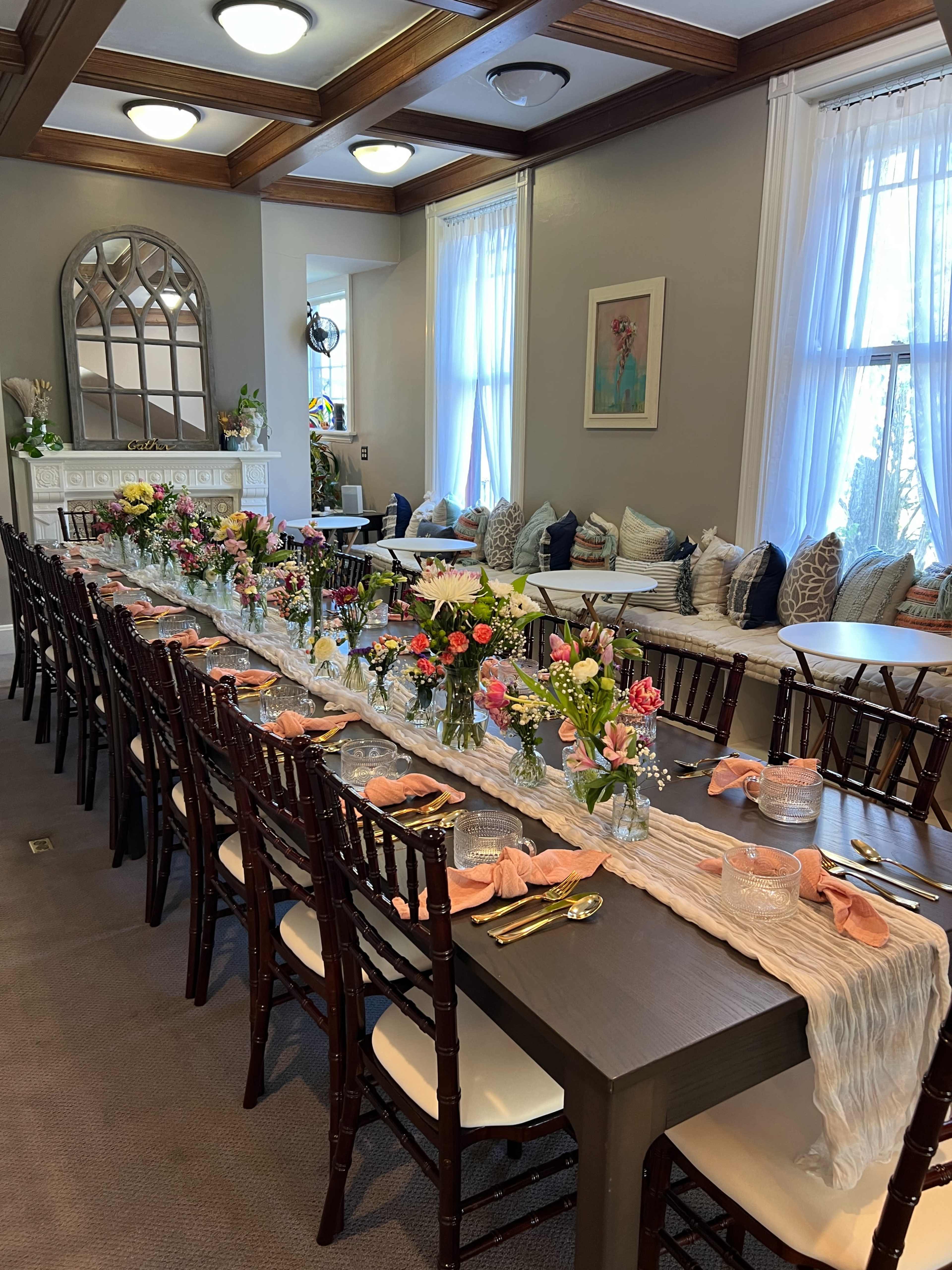 A long dining table is set with floral arrangements and decorative tableware in a brightly lit room featuring a fireplace and cushioned seating.