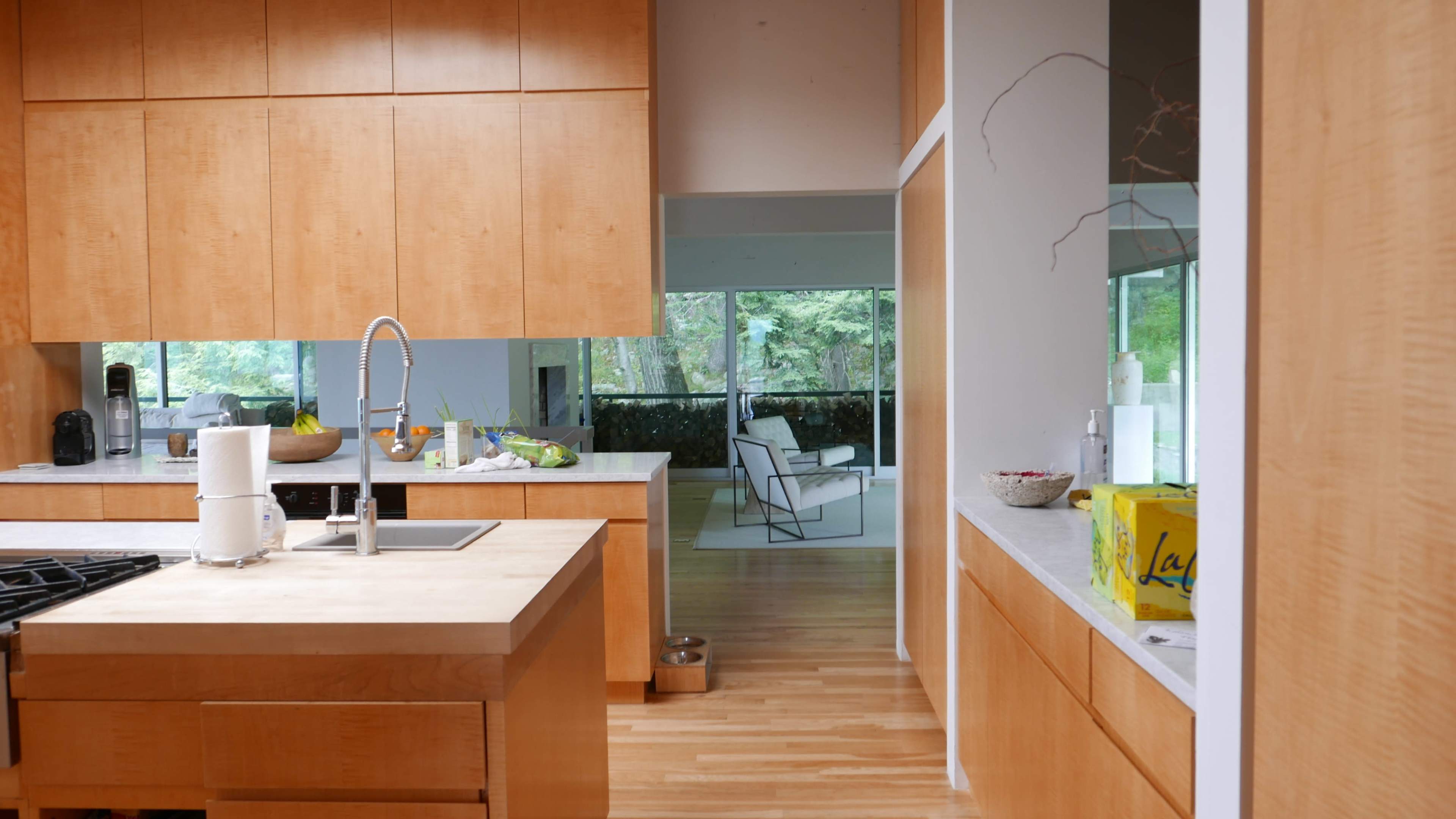 A modern kitchen with wooden cabinetry, a central island with a sink, and a view into a living area with large windows.