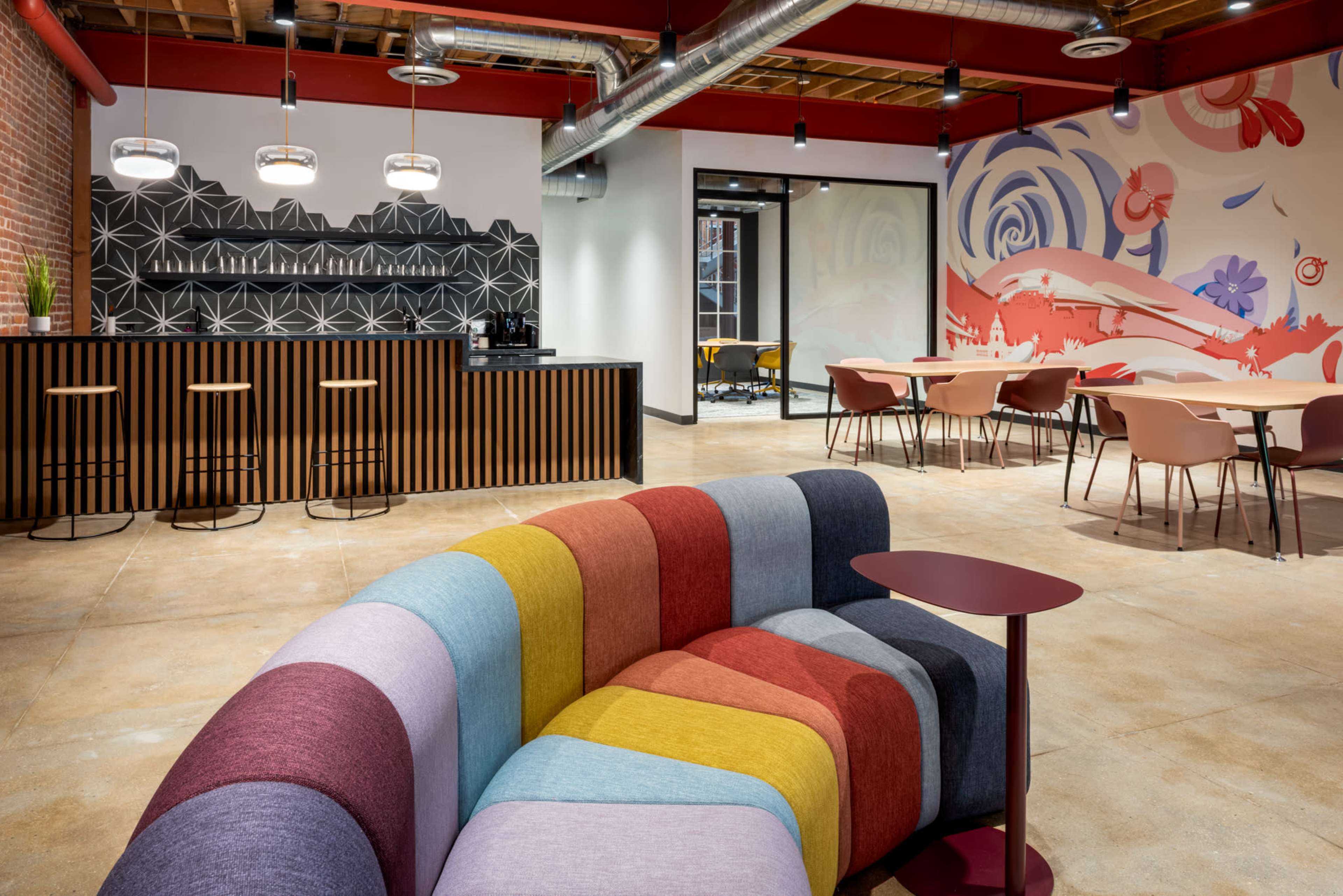The image shows a modern office interior featuring a colorful fabric couch, a bar with high stools, and tables arranged in a collaborative workspace.