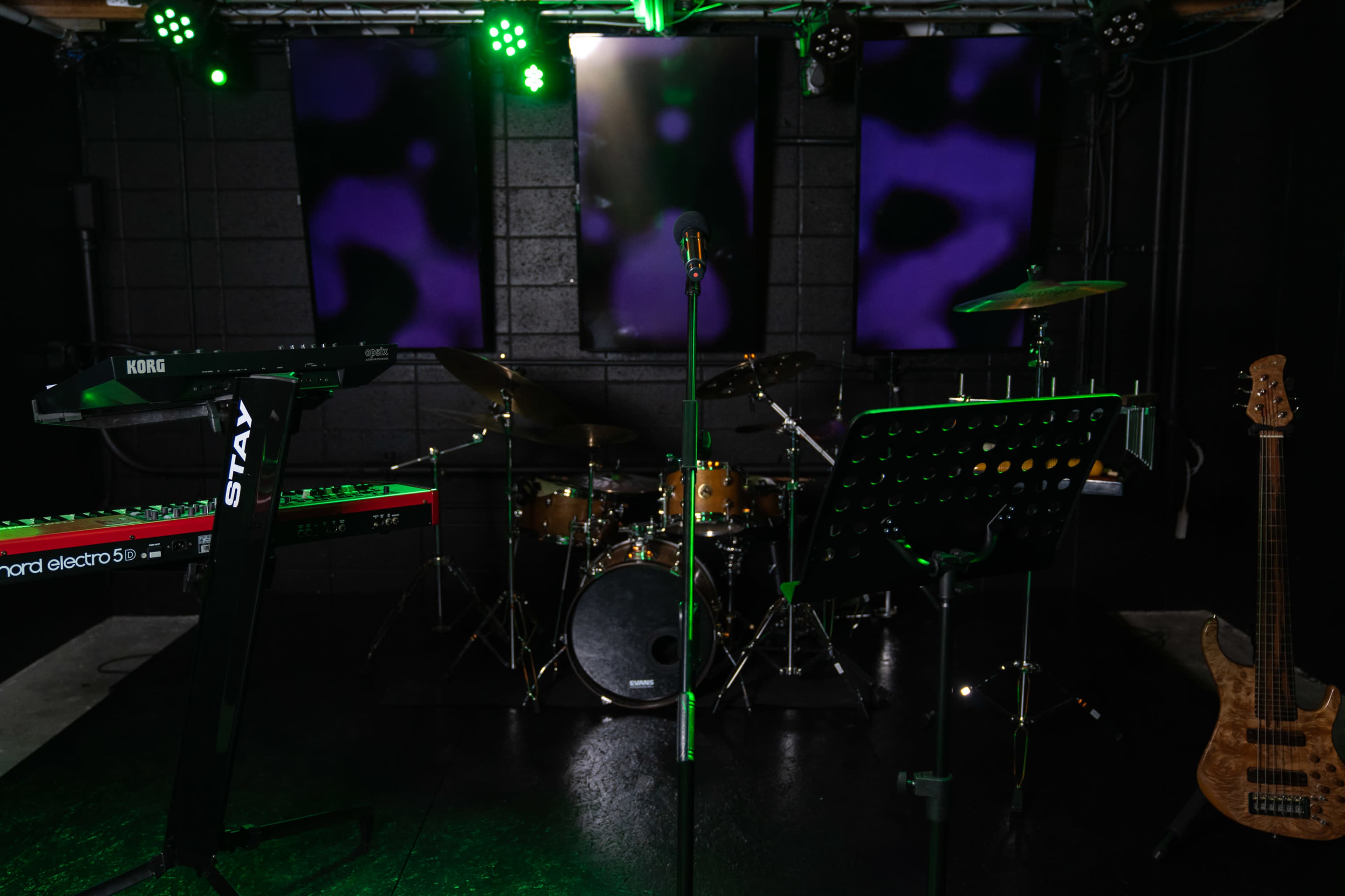 A music performance setup with a microphone in front of a drum kit, keyboard, and bass guitar, all under green stage lights and illuminated backdrops.