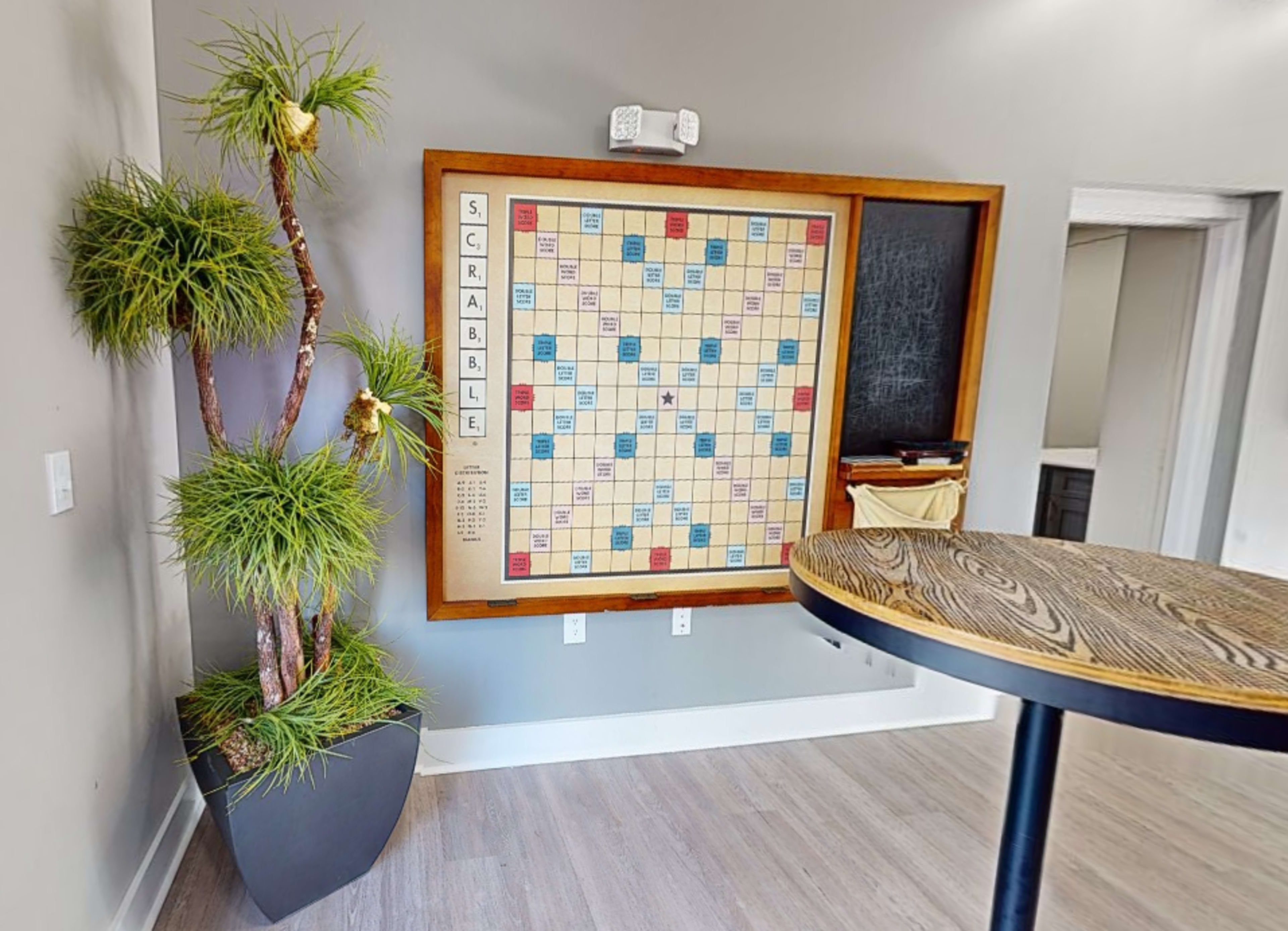 The image shows a Scrabble board mounted on the wall next to a tall potted plant in a modern room with wooden flooring.