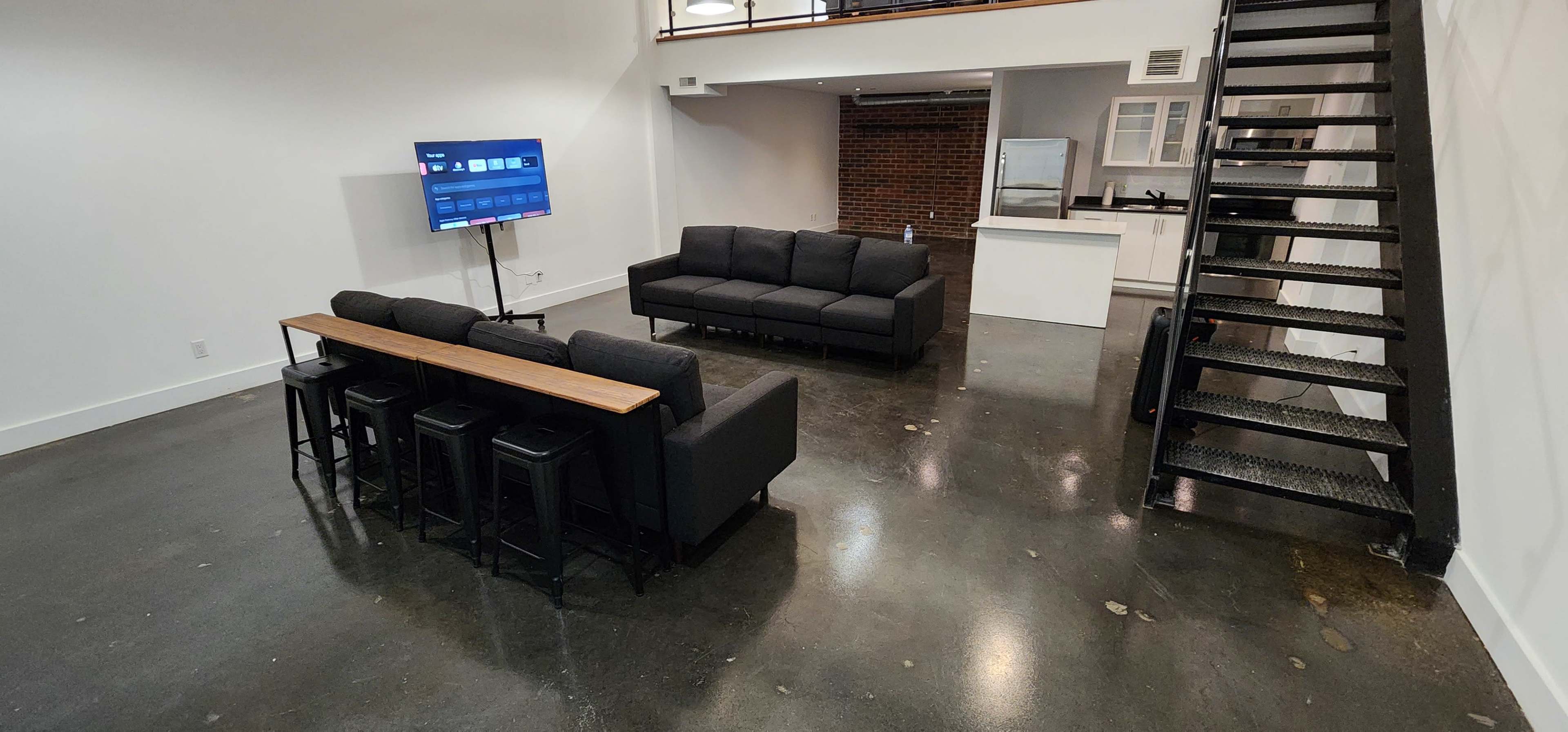 A spacious living area with a black sofa set facing a television, and a dining table with stools behind it, while a staircase leads to an upper level.